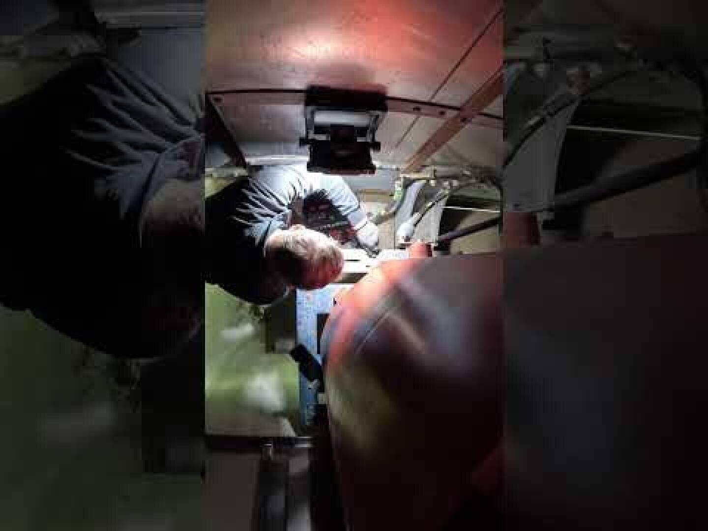 Eine Person arbeitet an einer Maschine in einem begrenzten Raum und benutzt Werkzeuge bei schwacher Beleuchtung. Die Person greift nach Rohren und mechanischen Bauteilen, in deren Nähe ein roter Tank zu sehen ist.