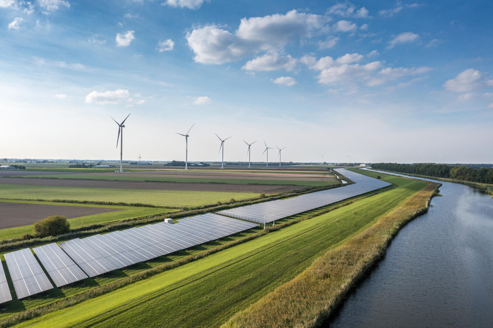 Aerial view of solar panels and wind turbines in a green landscape with a canal running alongside fields under a blue sky with scattered clouds.