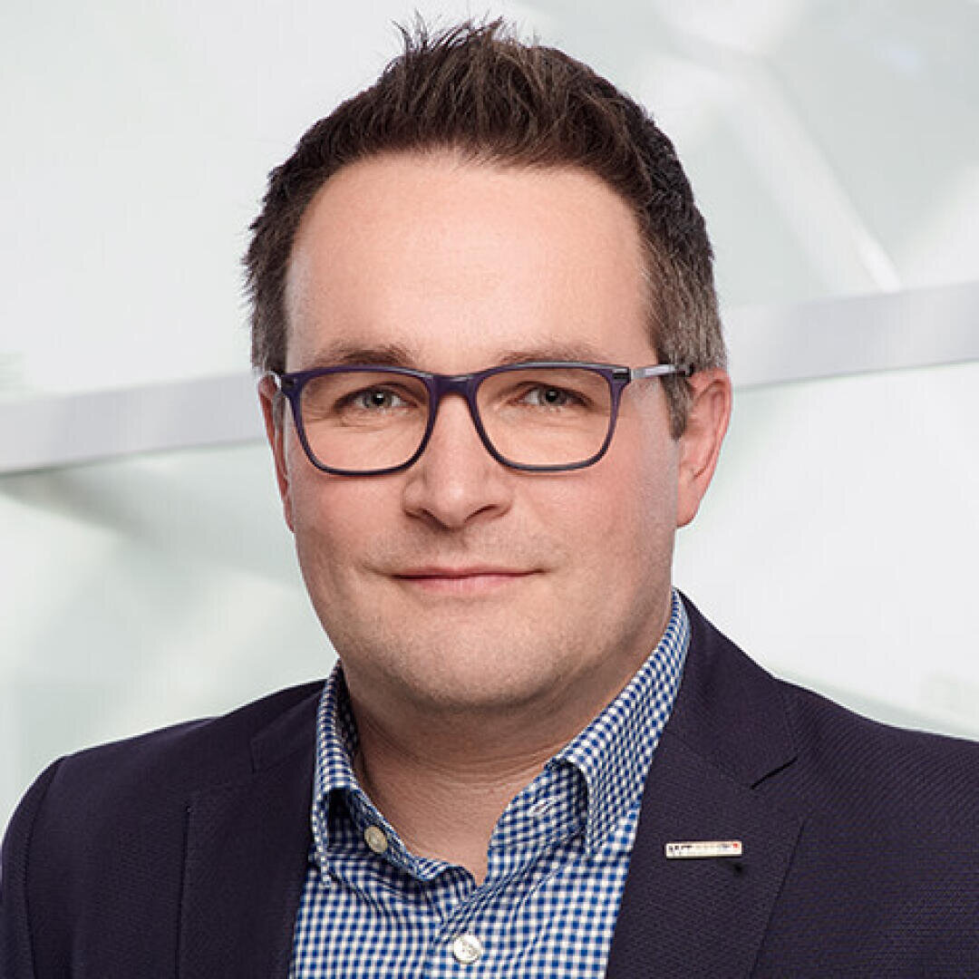 Ein Mann mit kurzen braunen Haaren und Brille, der einen dunklen Blazer über einem blau-karierten Hemd trägt, lächelt vor einem hellen, unscharfen Hintergrund freundlich in die Kamera.