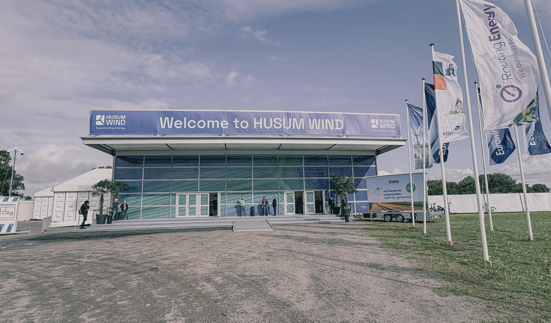 Ein großes Gebäude mit einem Schild mit der Aufschrift Willkommen bei HUSUM WIND am Eingang. Mehrere Personen stehen in der Nähe der Türen, und auf der rechten Seite sind große Fahnen mit verschiedenen Logos angebracht. Der Himmel ist teilweise bewölkt.