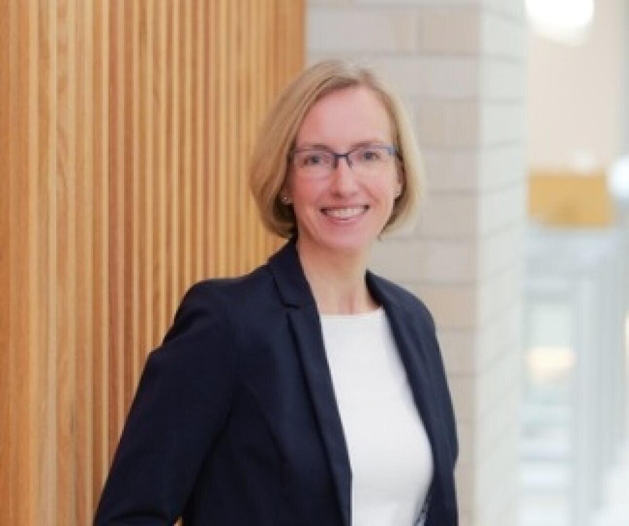 A woman with short blonde hair and glasses, dressed in a dark blazer and white top, smiles while standing indoors near a wooden panel wall and a white brick background.