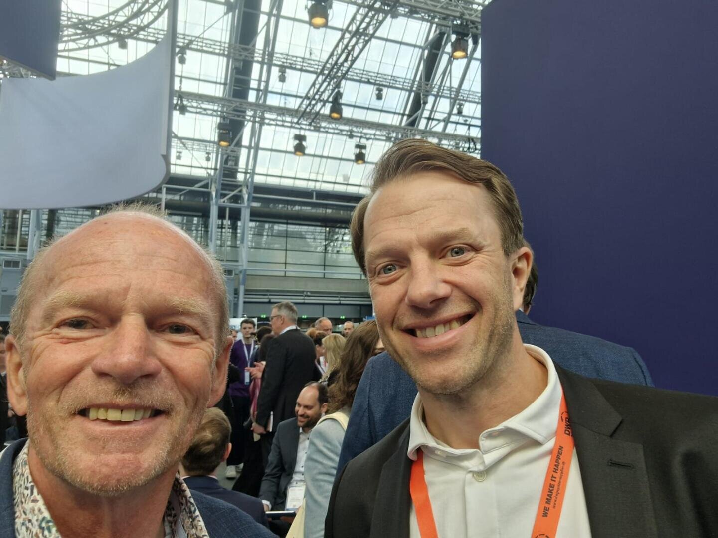 Two smiling men take a selfie at a busy indoor event with people in the background. The venue has a glass ceiling and modern metal scaffolding. One man is wearing a flowered shirt, the other a white shirt and an orange lanyard.