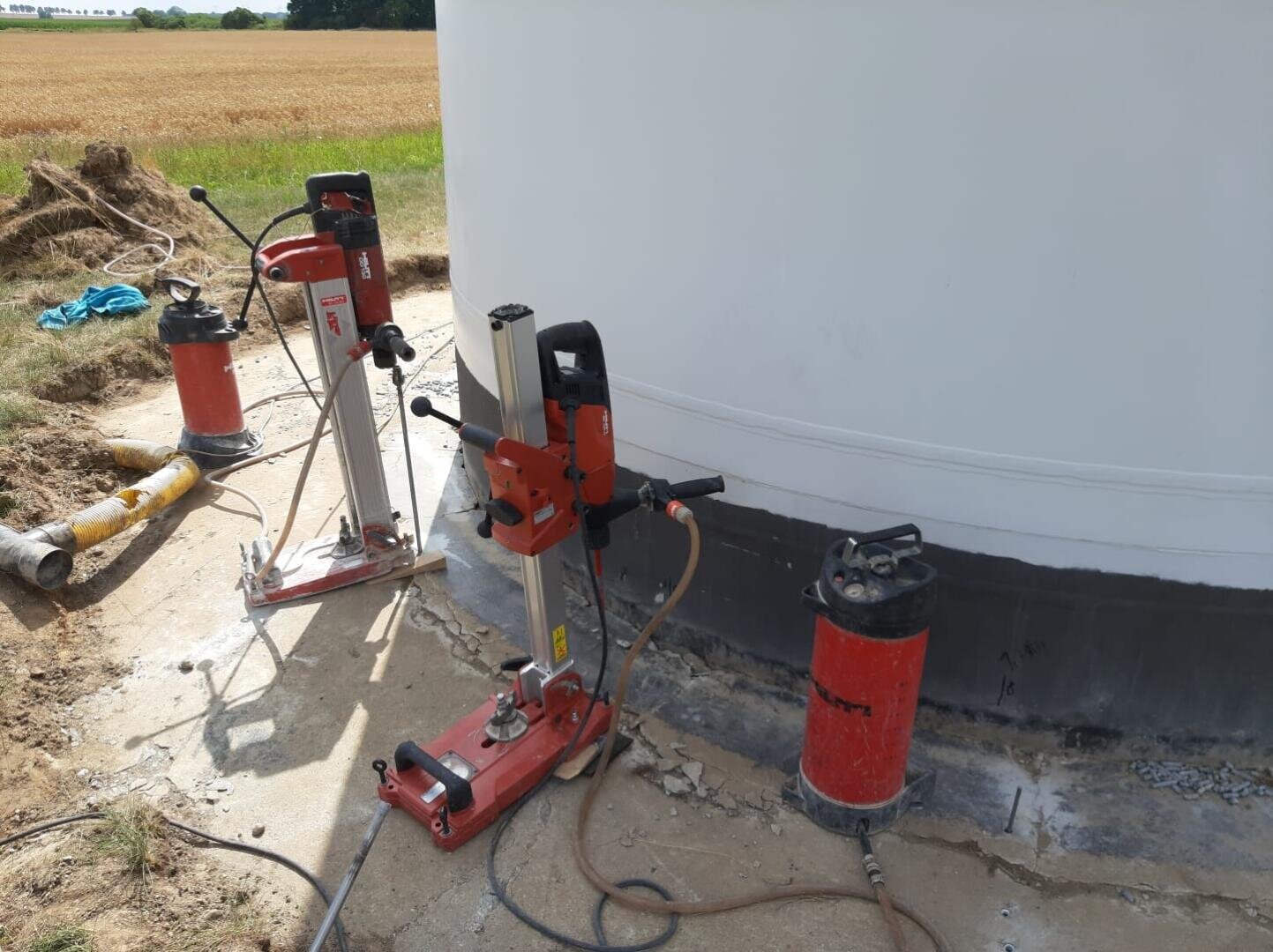 Drei rote und silberne Kernbohrmaschinen stehen auf dem Betonsockel eines großen weißen Gebäudes im Freien, in der Nähe eines grasbewachsenen Feldes und von Feldern unter einem sonnigen Himmel.