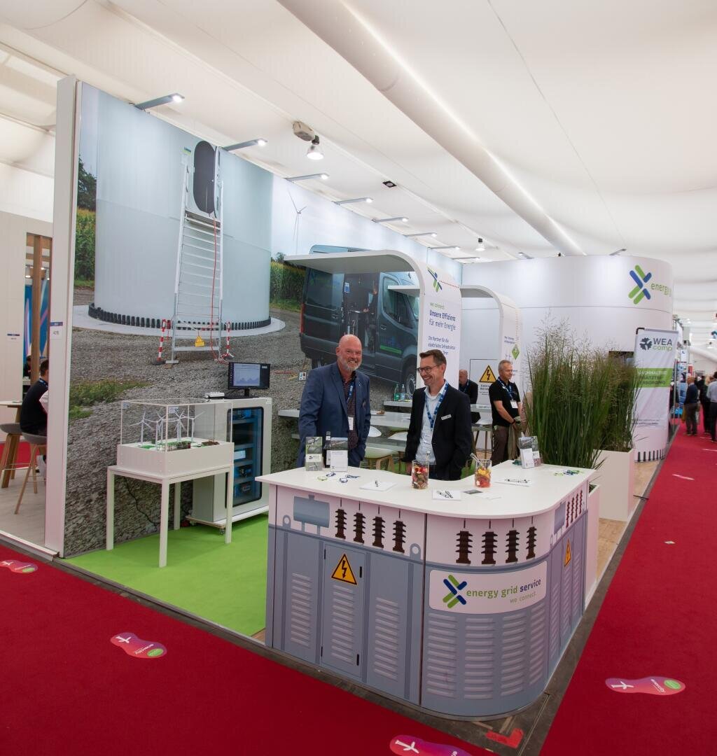 Two men in suits talk at a booth with branded energy grid services, systems and equipment displays. In the background, other people and booths can be seen on a red carpet.