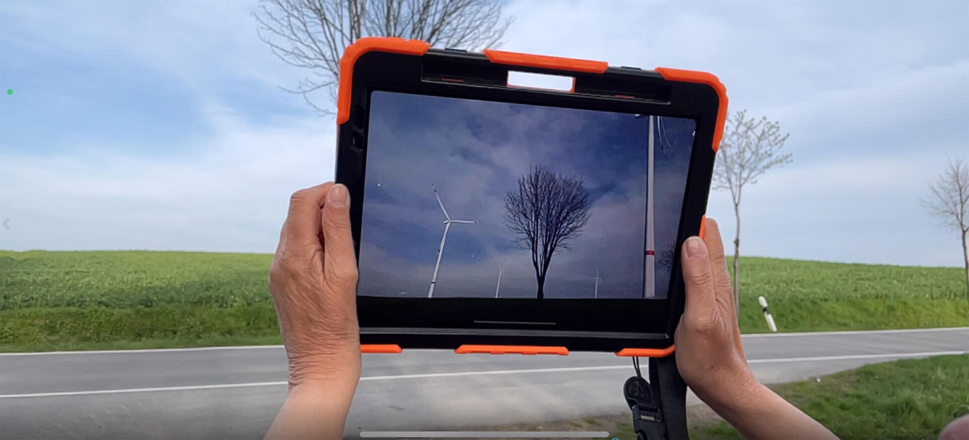 Eine Person hält ein Tablet in der Hand und fotografiert oder filmt Windturbinen und einen Baum in einer grasbewachsenen Landschaft am Straßenrand; auf dem Bildschirm des Tablets ist die Szene mit einem anderen Himmel zu sehen.