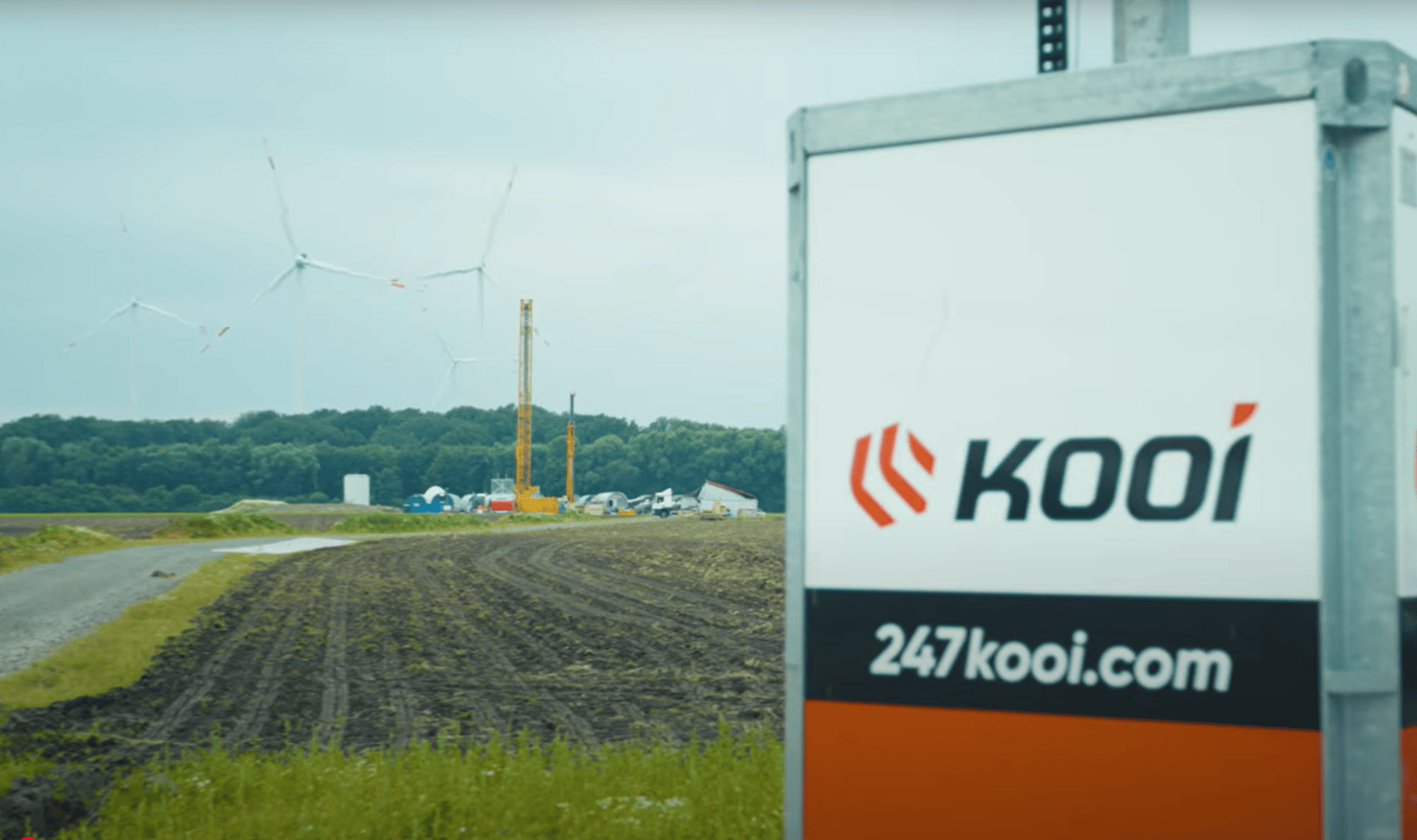 A sign with the Kooi logo and the website 247kooi.com stands in the foreground of a construction site with wind turbines, cranes and trees in the background under a cloudy sky.
