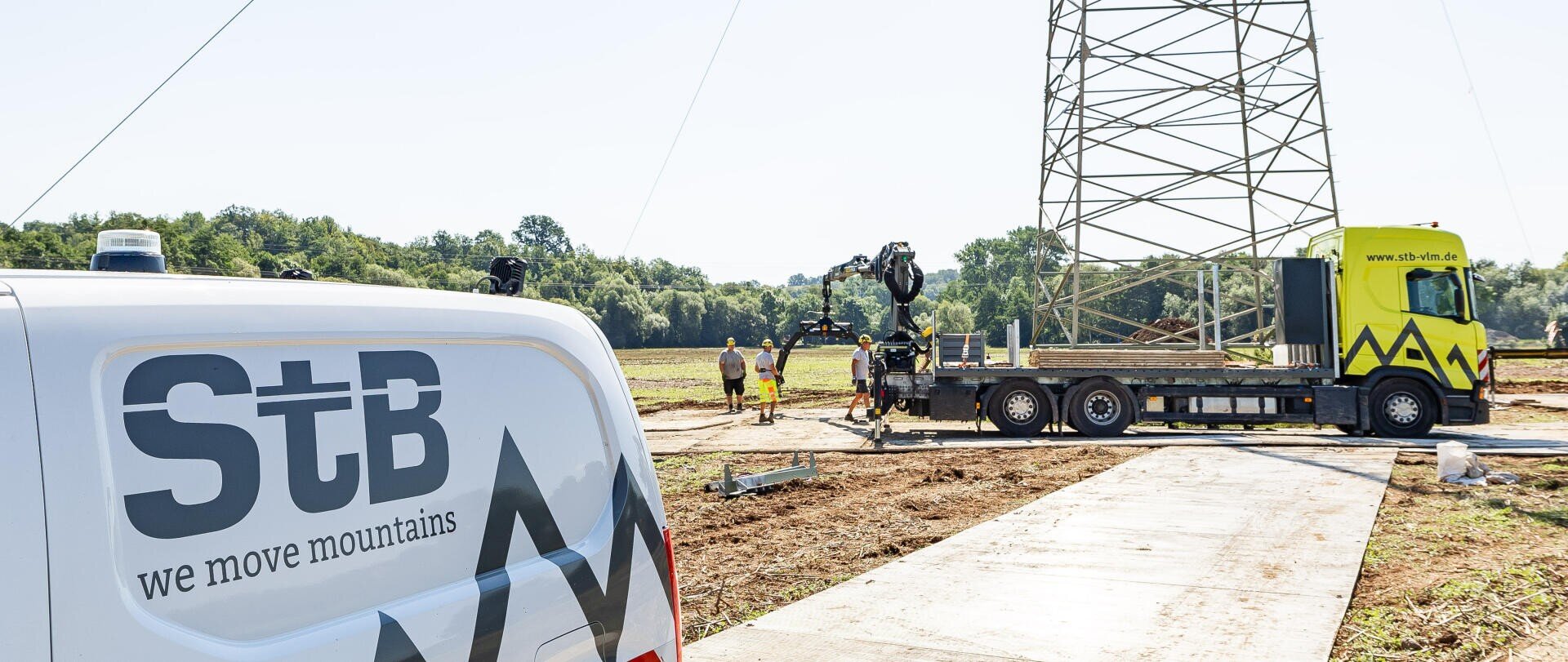 Ein weißer Lieferwagen mit StB wir versetzen Berge parkt in der Nähe einer Baustelle, wo Arbeiter und ein gelber LKW mit einem Kran an einem großen Metallturm auf einem offenen Feld stehen.