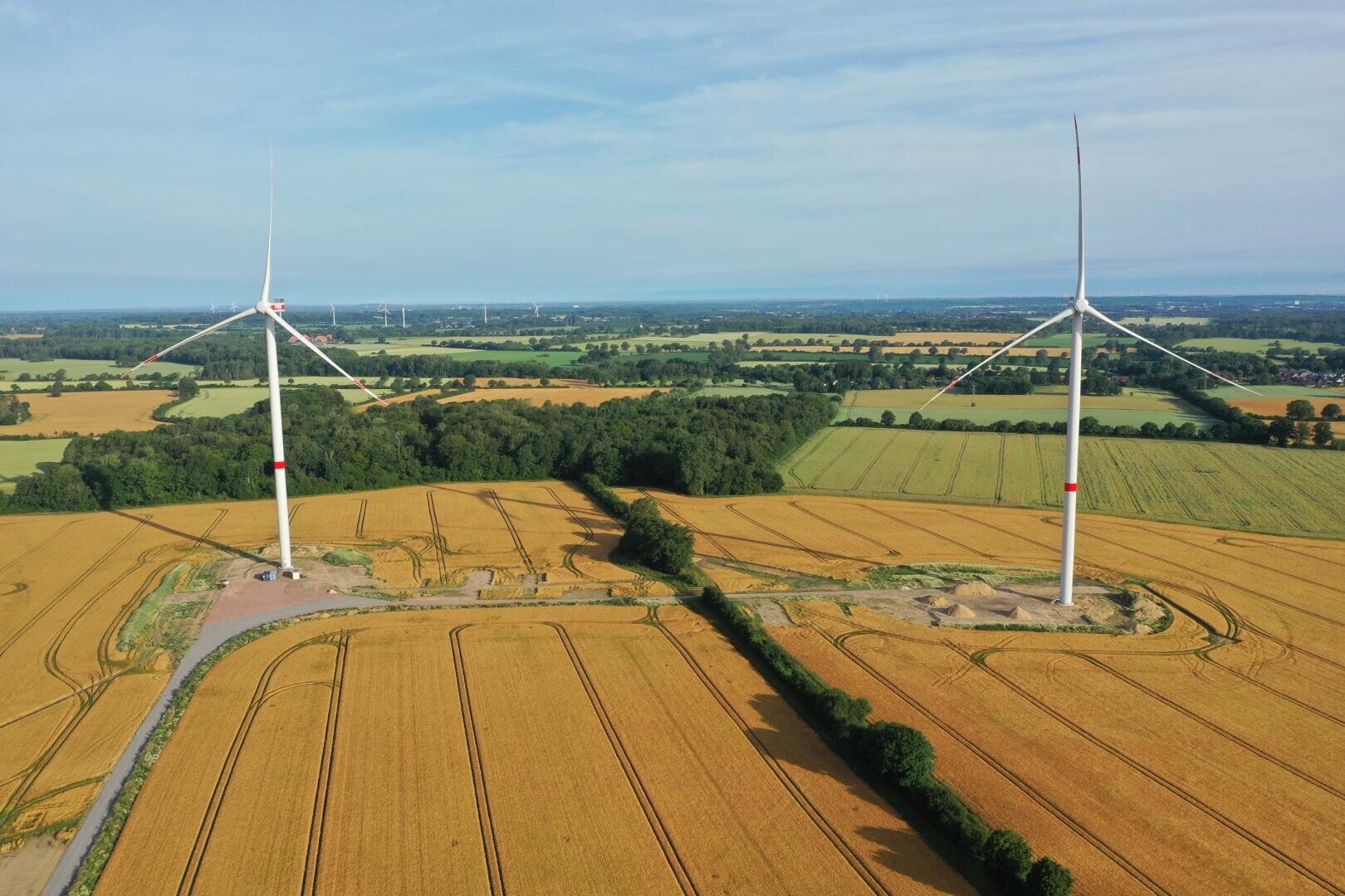 Luftaufnahme von zwei großen Windturbinen, die in goldenen Feldern stehen, umgeben von grünen Bäumen und Ackerland unter einem blauen Himmel.