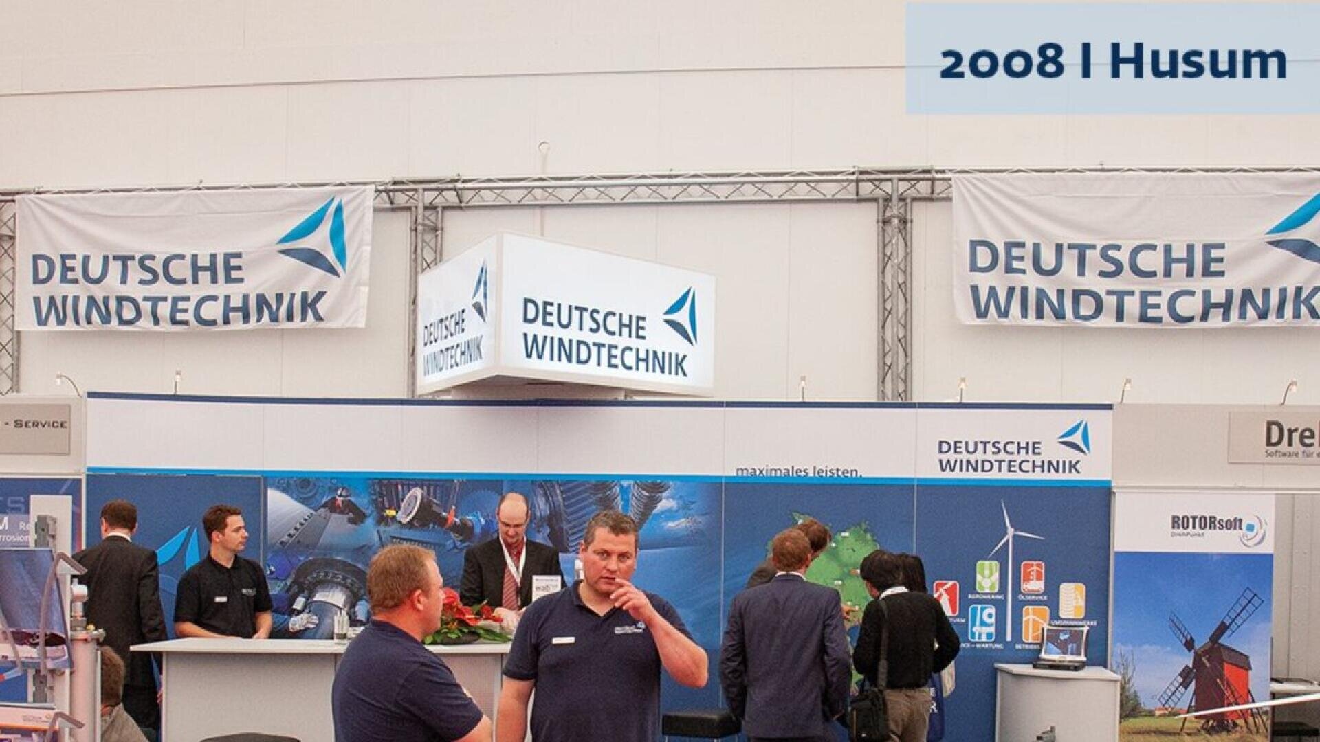 Booth for Deutsche Windtechnik at a trade show, featuring banners, displays about wind energy, and people talking. A sign in the corner reads 2008 | Husum.