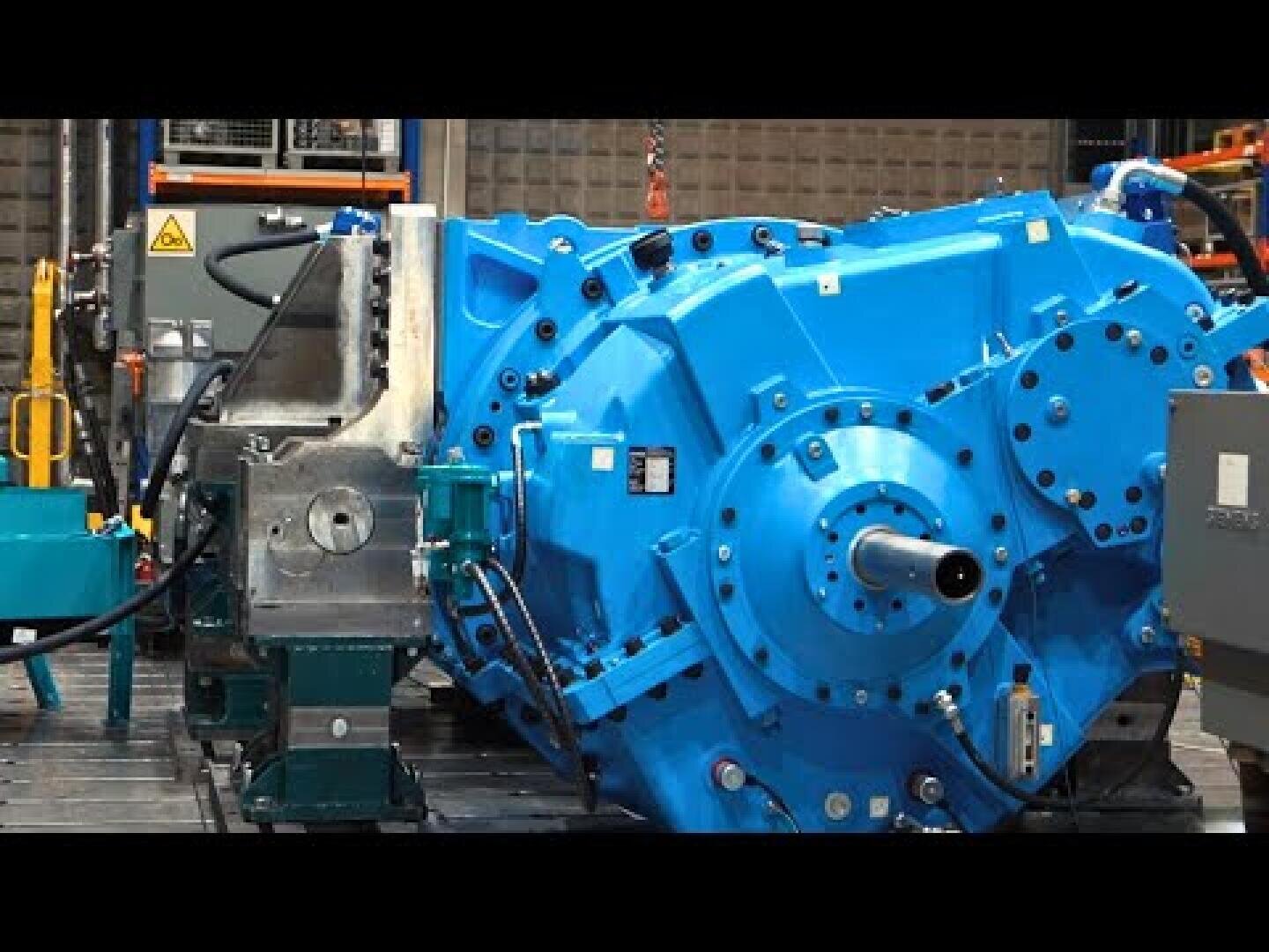 Large industrial blue gearbox with multiple bolts and a cylindrical shaft, set in a workshop environment with various machinery and equipment visible in the background.