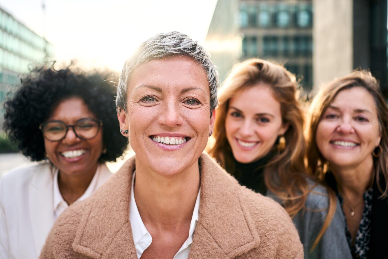 Vier Frauen stehen im Freien dicht beieinander und lächeln in die Kamera. Das Sonnenlicht scheint hinter ihnen und schafft eine warme und fröhliche Atmosphäre. Sie wirken fröhlich und freundlich, gekleidet in Freizeit- und Geschäftskleidung.