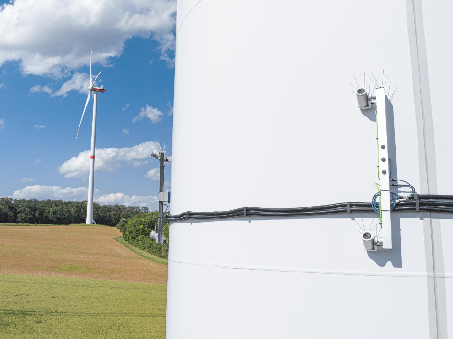 A close-up of a wind turbine tower with visible cables and small devices attached, standing in a green field. Another wind turbine is seen in the background under a partly cloudy blue sky.