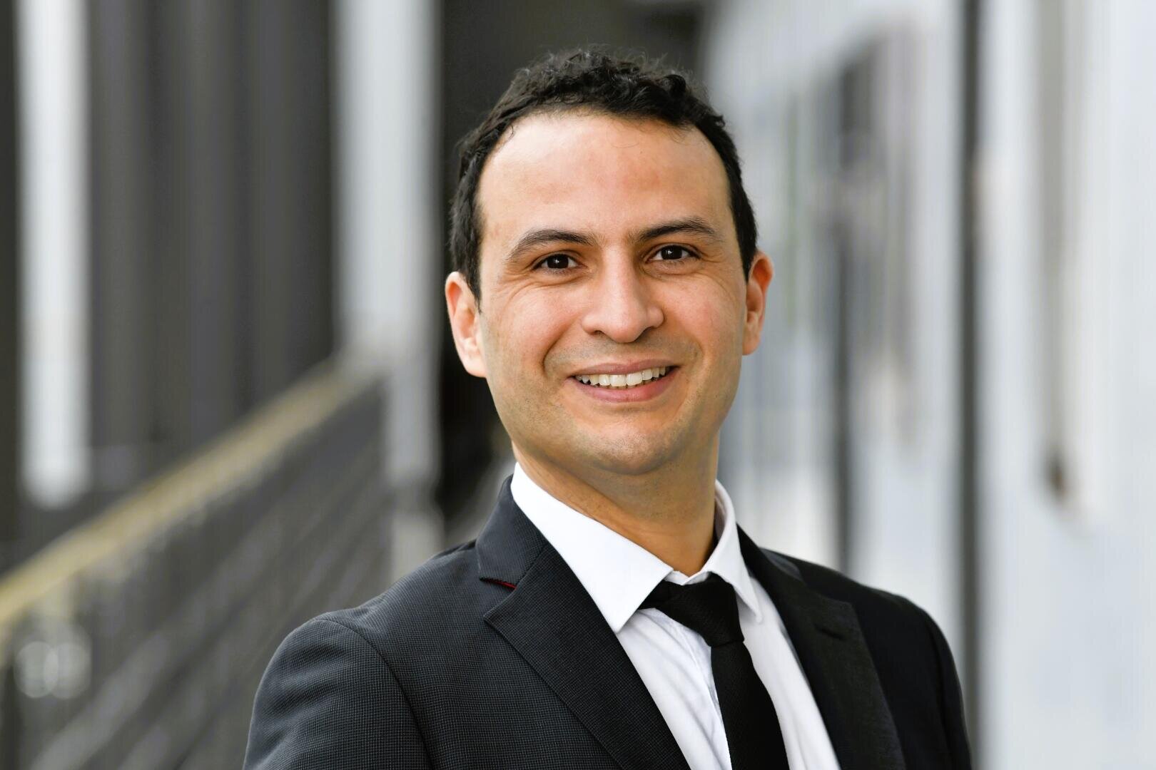 Ein Mann in Anzug und Krawatte lächelt in die Kamera in einem Innenraum mit unscharfen Säulen und Mauern im Hintergrund.