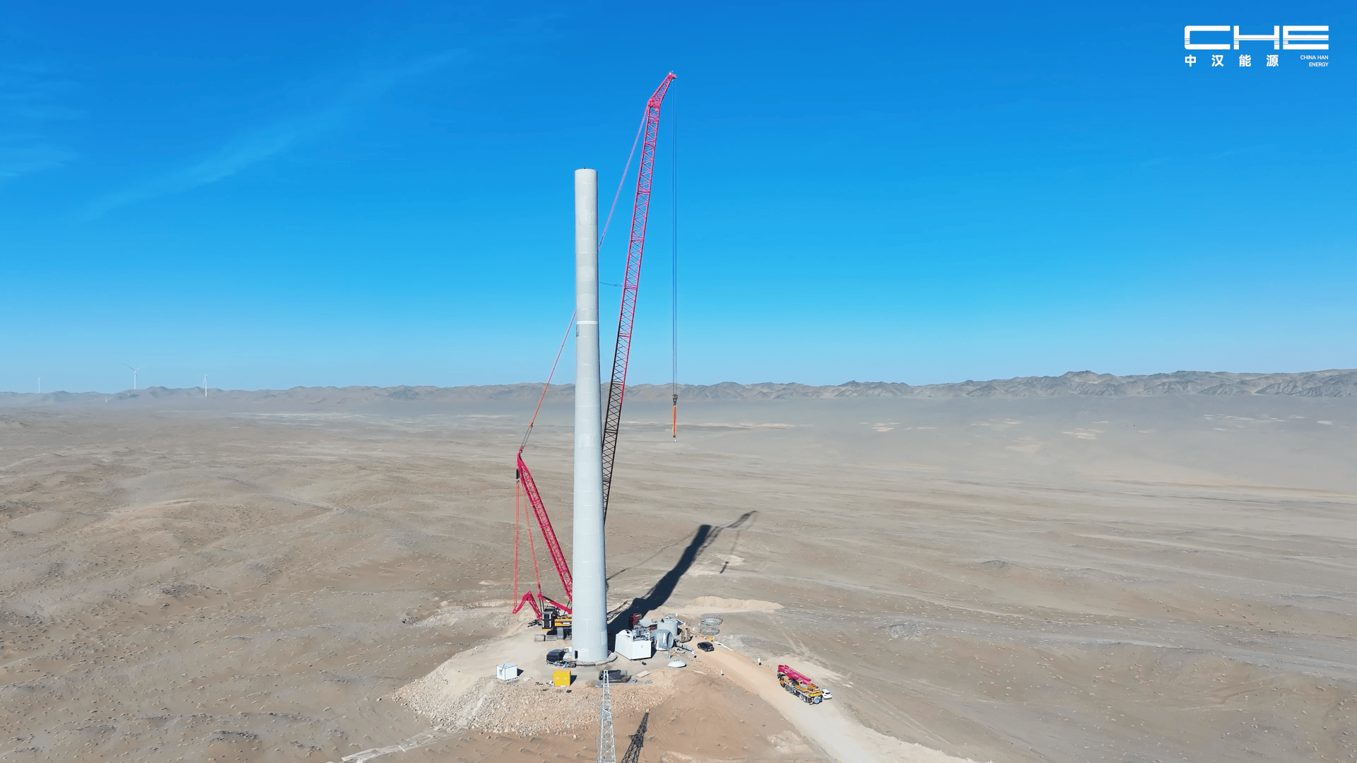 A tall wind turbine tower under construction in a barren desert landscape, next to it two red cranes and some vehicles on the ground; mountains can be seen in the distant background.