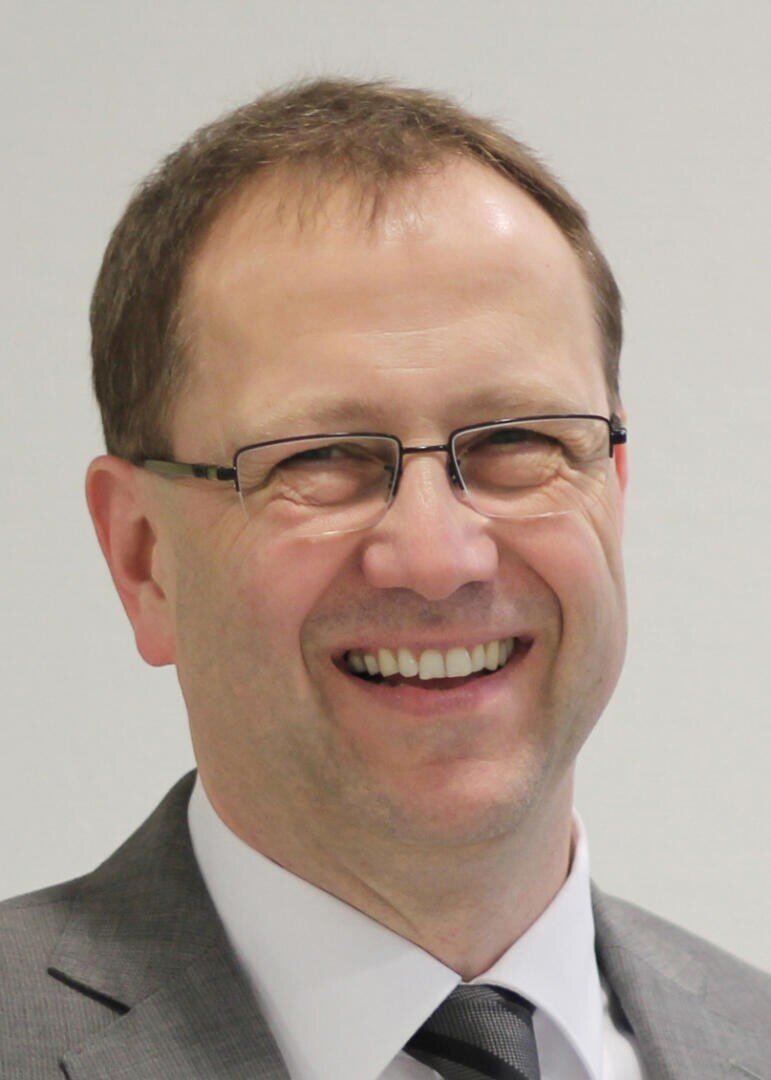 Ein Mann mittleren Alters mit kurzen braunen Haaren, der eine Brille und einen grauen Anzug mit weißem Hemd und dunkler Krawatte trägt, lächelt vor einem schlichten hellen Hintergrund.
