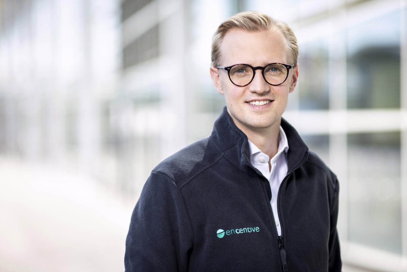 Ein lächelnder Mann mit blondem Haar und Brille, der eine dunkle Jacke mit dem enCentive-Logo trägt, steht vor einem verschwommenen modernen Glasgebäude.