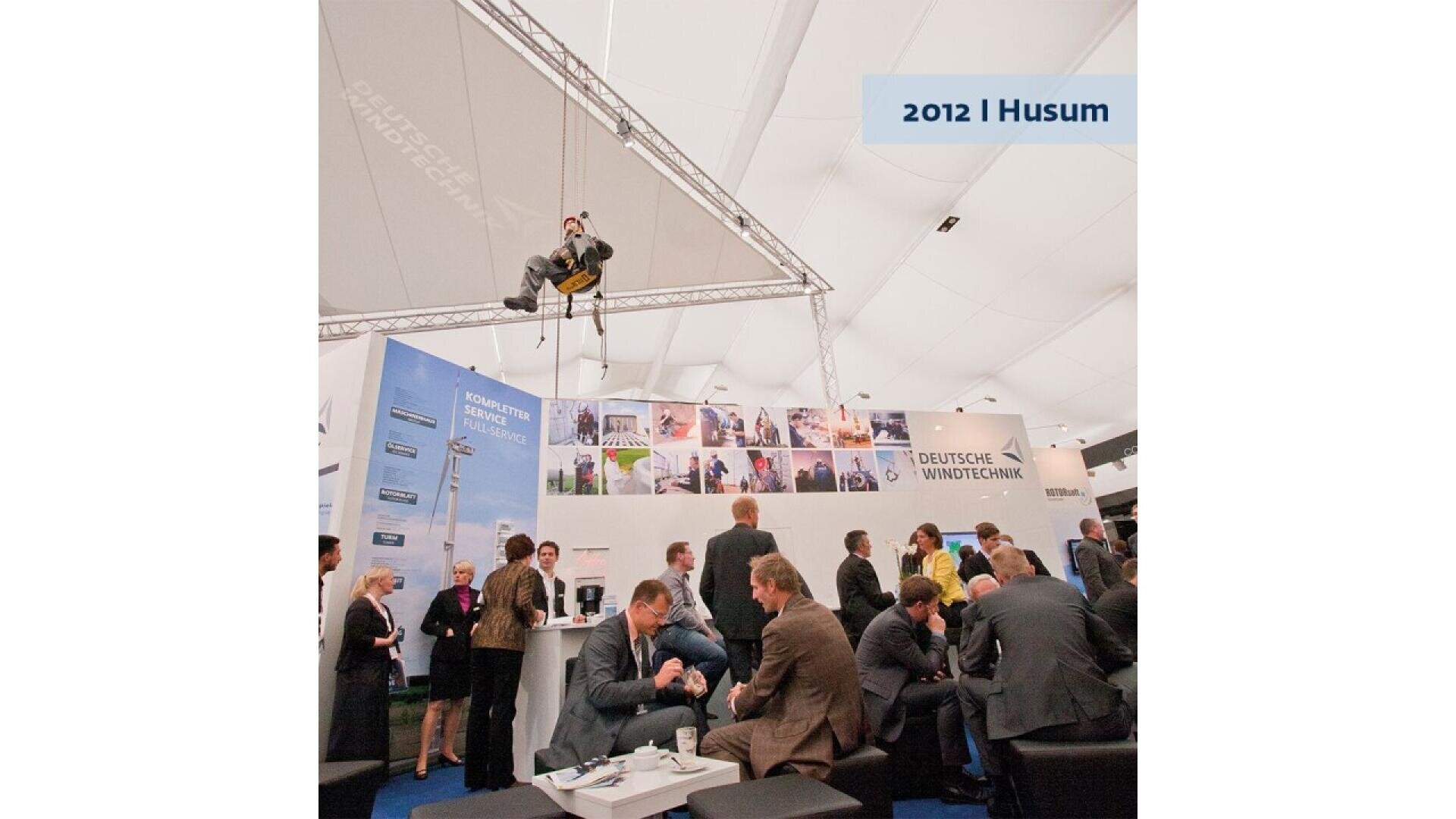A person is suspended from the ceiling above a busy exhibition booth labeled Deutsche Windtechnik at the 2012 Husum event, while many people in business attire interact below.