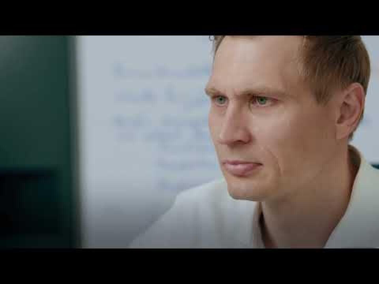 A man with short light brown hair and a serious expression is looking slightly to the side. He sits indoors with a blurred whiteboard or chart in the background.