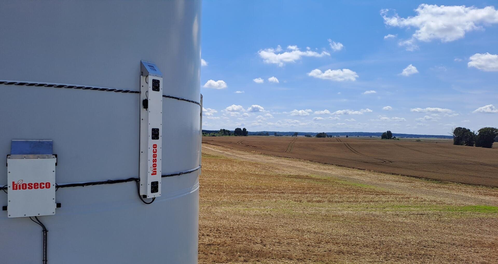 Nahaufnahme eines Überwachungsgeräts mit der Aufschrift bioseco, das auf einem Turm einer Windkraftanlage montiert ist und ein abgeerntetes Feld unter blauem Himmel mit vereinzelten Wolken überblickt.