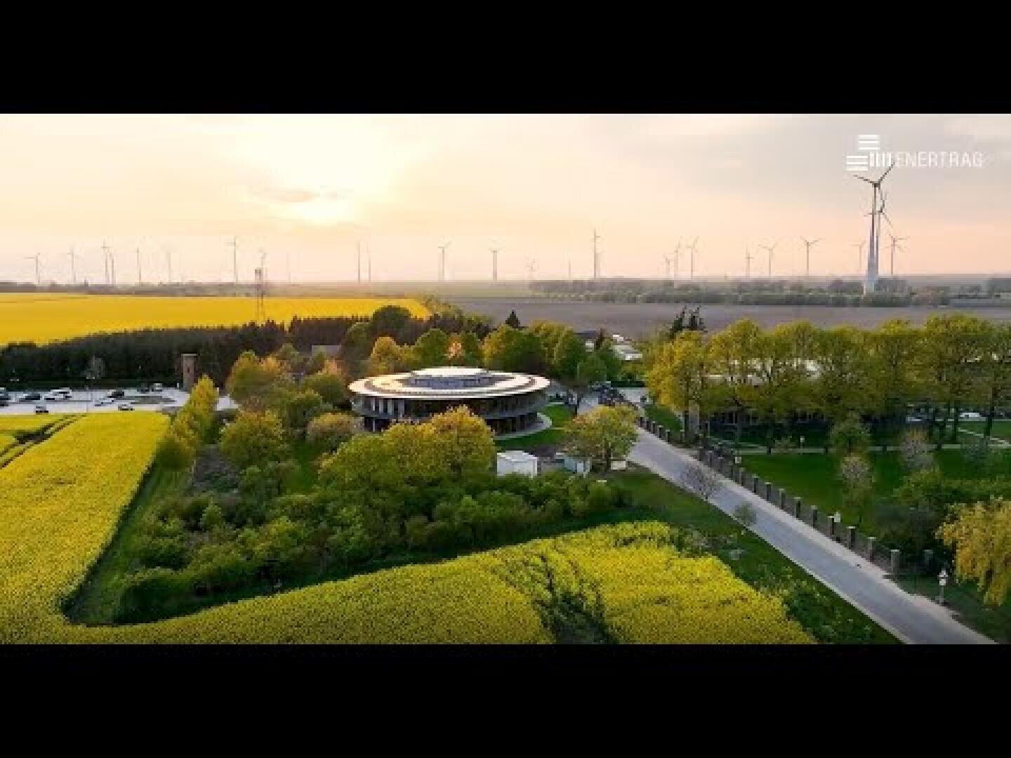 A circular building surrounded by trees and yellow fields, with wind turbines visible in the background under a hazy sky. The image has the Industria logo in the top right corner.