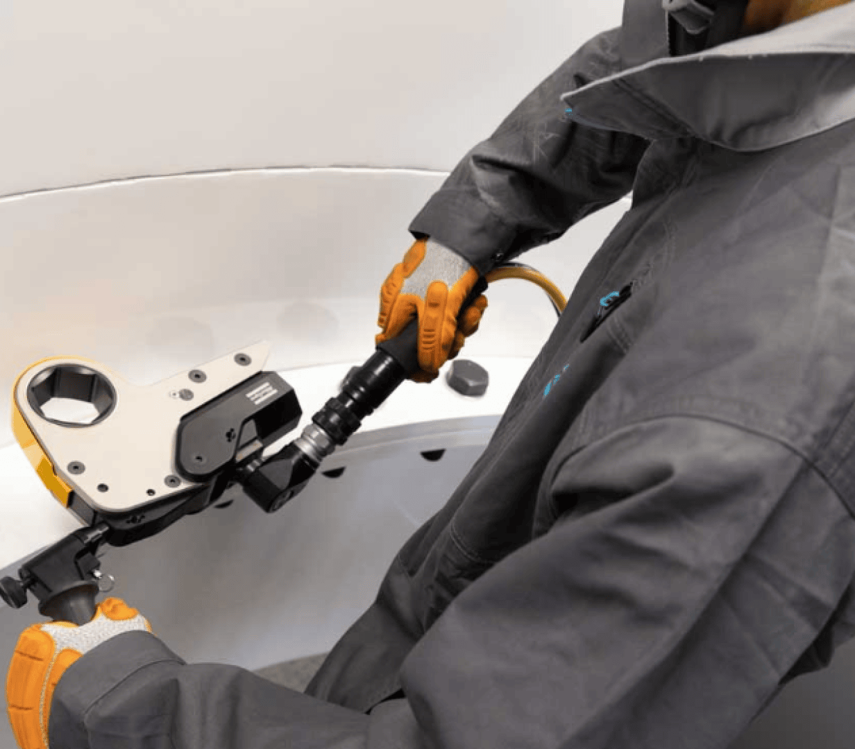 A person wearing gray protective clothing and orange gloves uses a hydraulic torque wrench to tighten a large bolt inside an industrial setting.