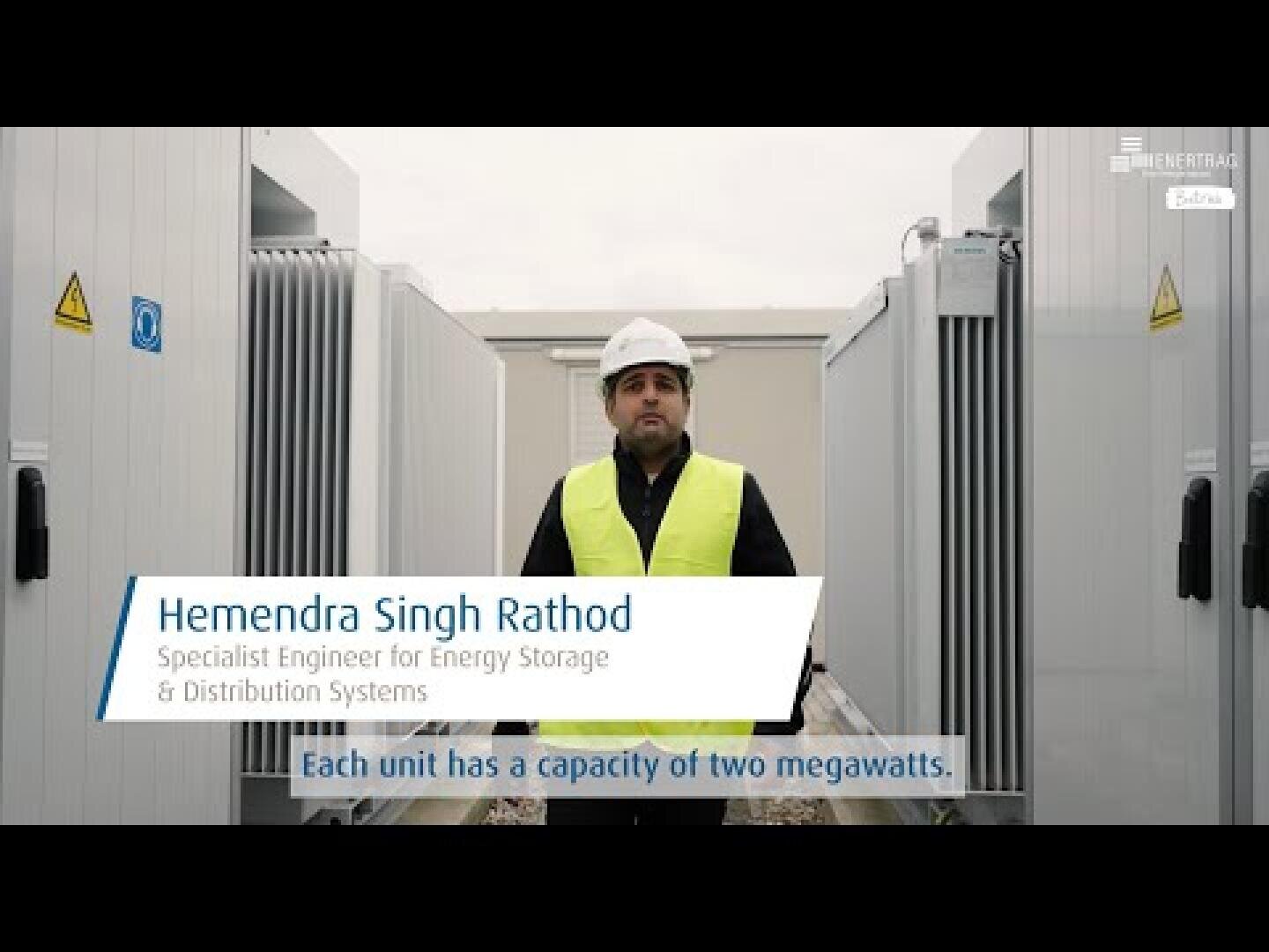 Ein Mann mit Schutzhelm und gelber Sicherheitsweste steht im Freien zwischen zwei großen Industrieanlagen. Der Text auf dem Bildschirm identifiziert ihn als Hemendra Singh Rathod, Fachingenieur für Energiespeicher- und Verteilungssysteme.