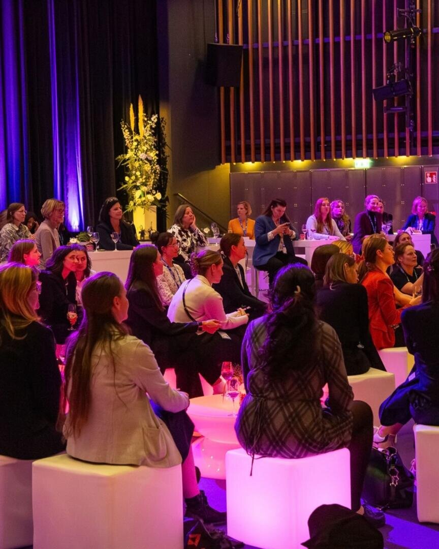 Bei einer Veranstaltung in einem Saal mit violetter Beleuchtung sitzen die Menschen auf leuchtenden Würfelsitzen. Einige blicken zu einem Redner oder einer Präsentation, während andere sich unterhalten. Im Hintergrund sind Blumen und Holzvertäfelungen zu sehen.