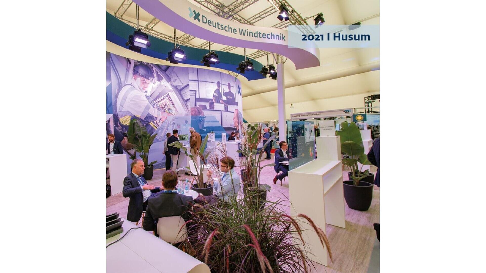 People sit and talk at a booth for Deutsche Windtechnik at an indoor expo. The booth features plants, large technology displays, and a curved sign overhead reading 2021 | Husum. Bright lights illuminate the area.