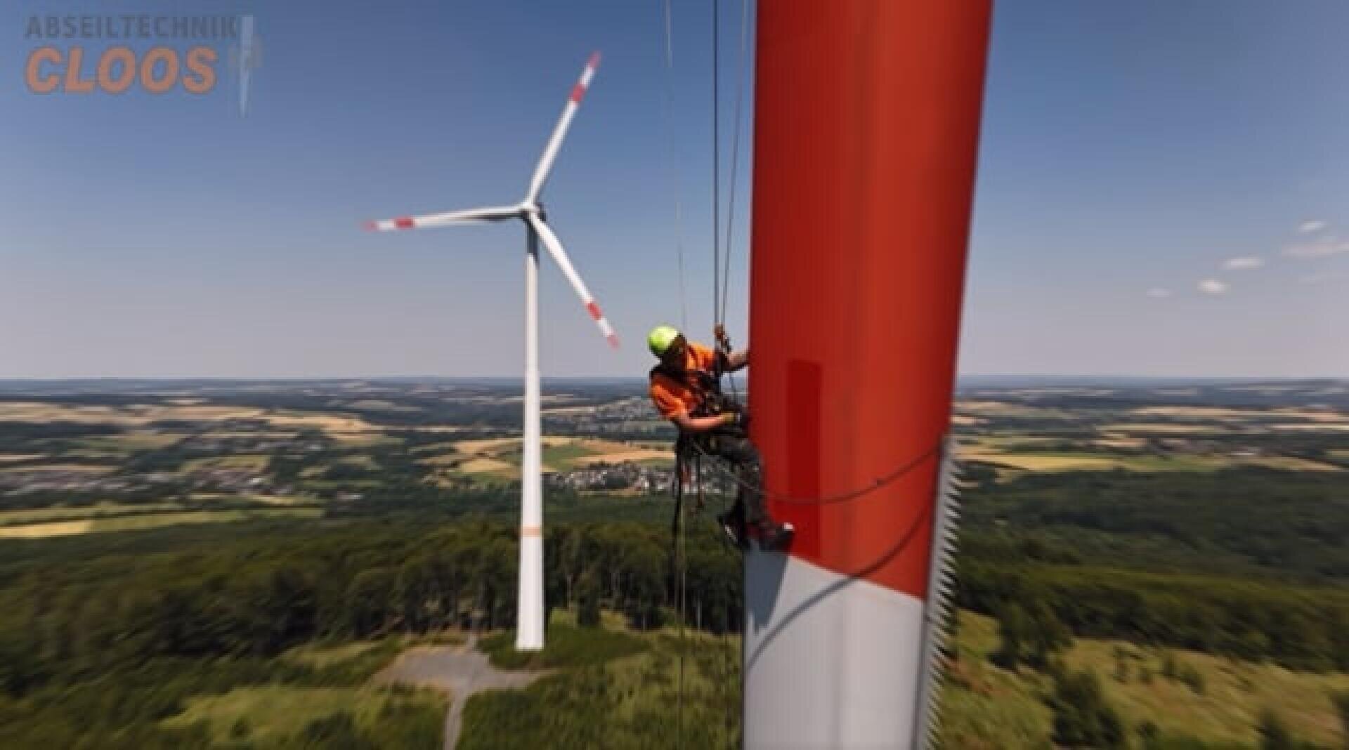 Ein Arbeiter in Schutzausrüstung ist an Seilen aufgehängt und inspiziert oder wartet eine große rot-weiße Windkraftanlage. Im Hintergrund steht eine weitere Windkraftanlage. Unter einem klaren Himmel sind Felder und Wälder zu sehen.