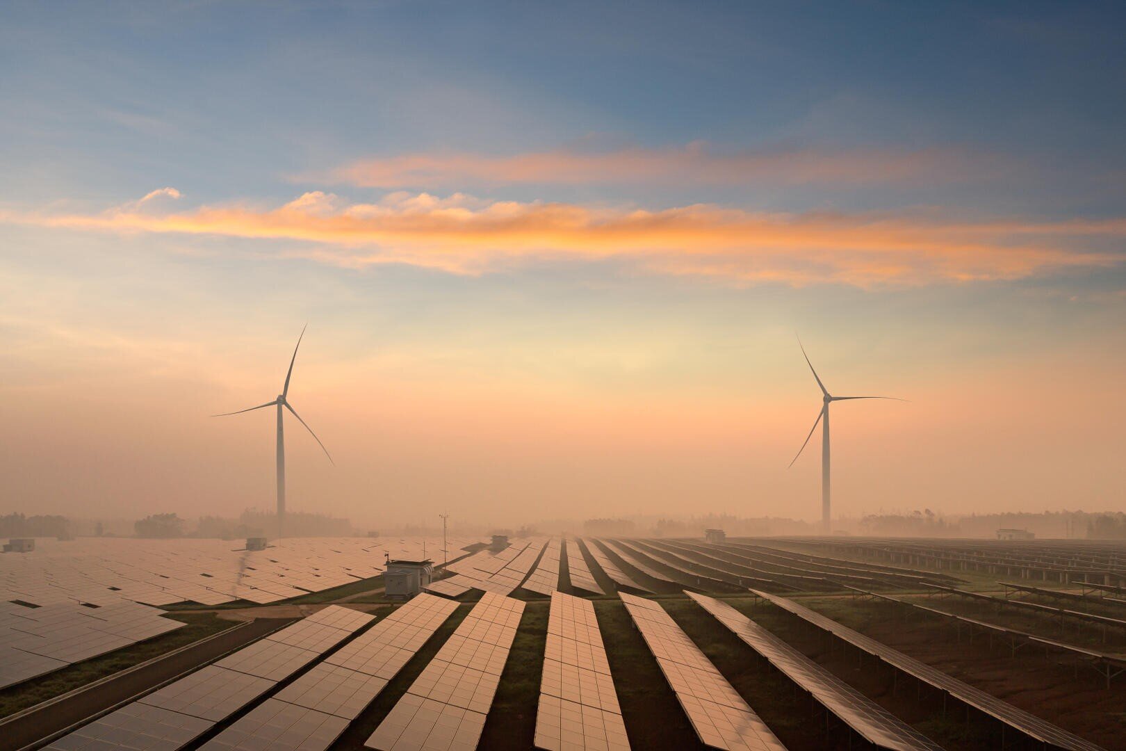 Rows of solar panels and two wind turbines stand on a large field at sunrise, with soft clouds and a warm orange sky in the background, symbolizing renewable energy sources.