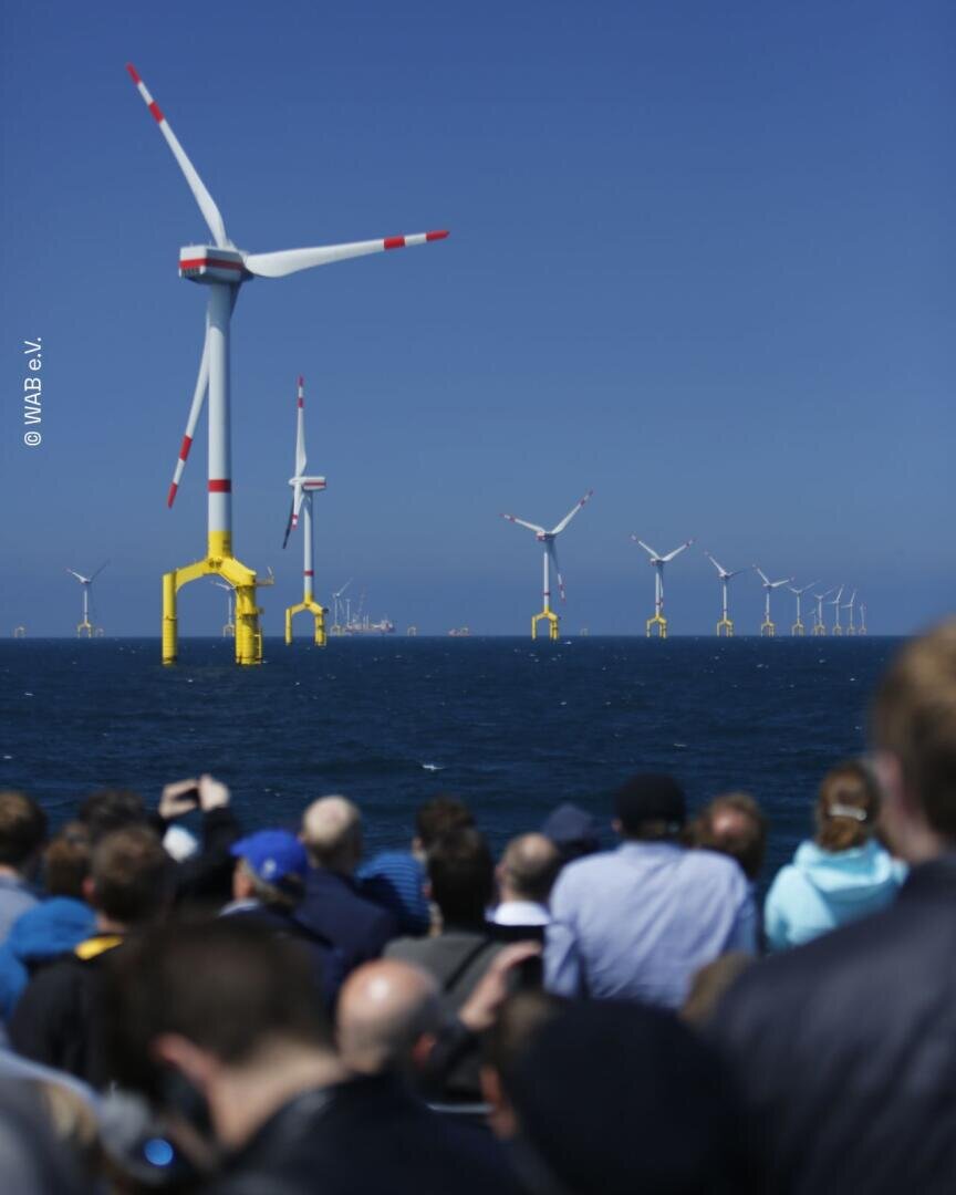 An einem klaren Tag, an dem der blaue Himmel und das blaue Wasser zu sehen sind, betrachtet eine Menschenmenge die im Meer aufgereihten Offshore-Windkraftanlagen. Die Anlagen stehen auf gelben Sockeln, und laut Textangabe links stammt das Foto von IWAB.