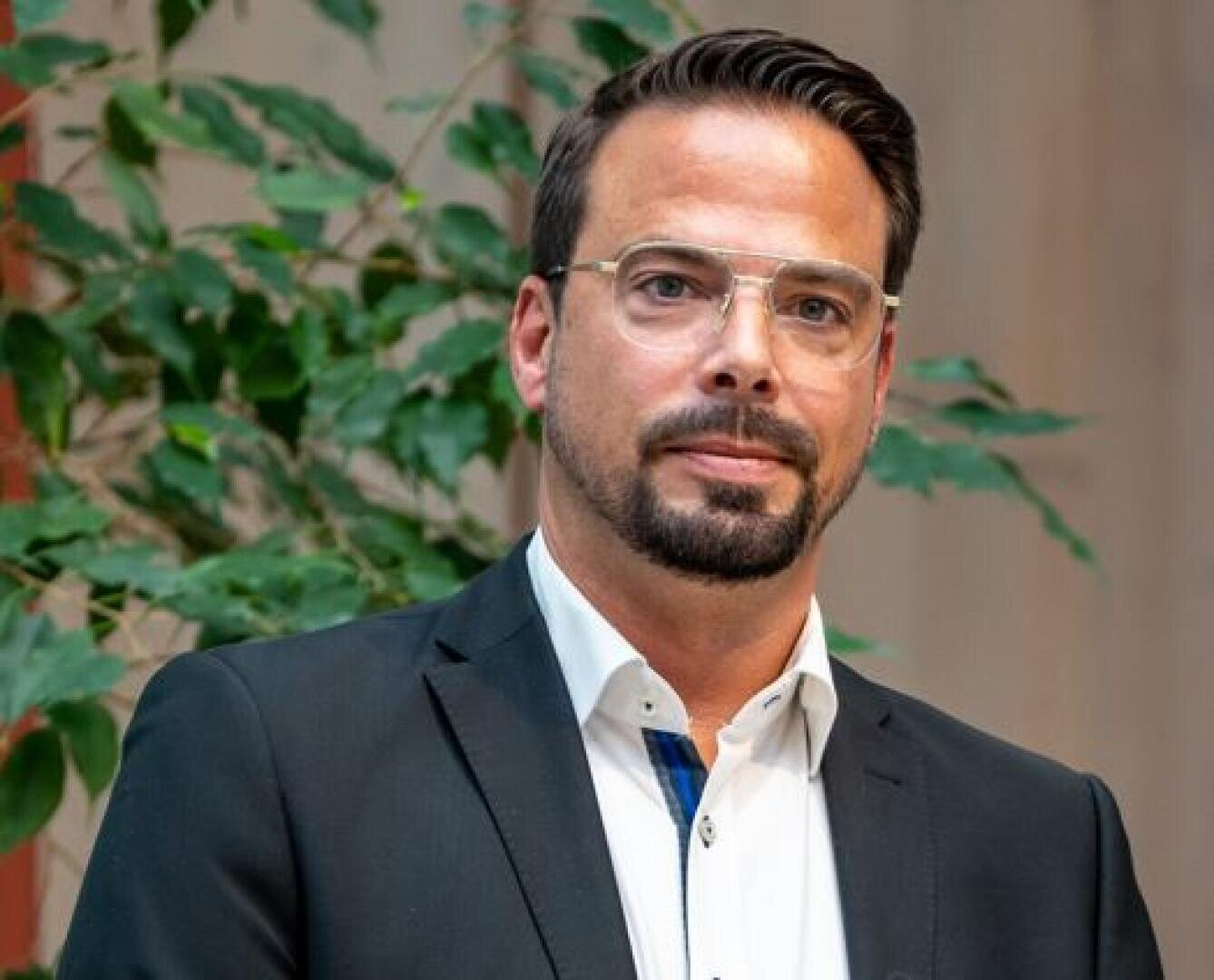 Ein Mann mit kurzen dunklen Haaren, einem gestutzten Bart und einer Brille trägt eine schwarze Anzugsjacke und ein weißes Hemd. Er steht in einem Innenraum vor einer grünen Blattpflanze.