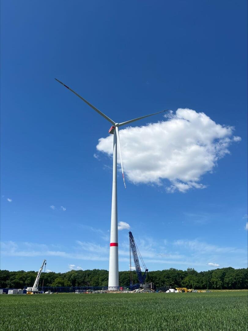 Eine hohe Windkraftanlage steht auf einer Wiese unter blauem Himmel mit einer einzelnen weißen Wolke. In der Nähe des Fundaments sind Baukräne und -geräte zu sehen, im Hintergrund eine Baumreihe.