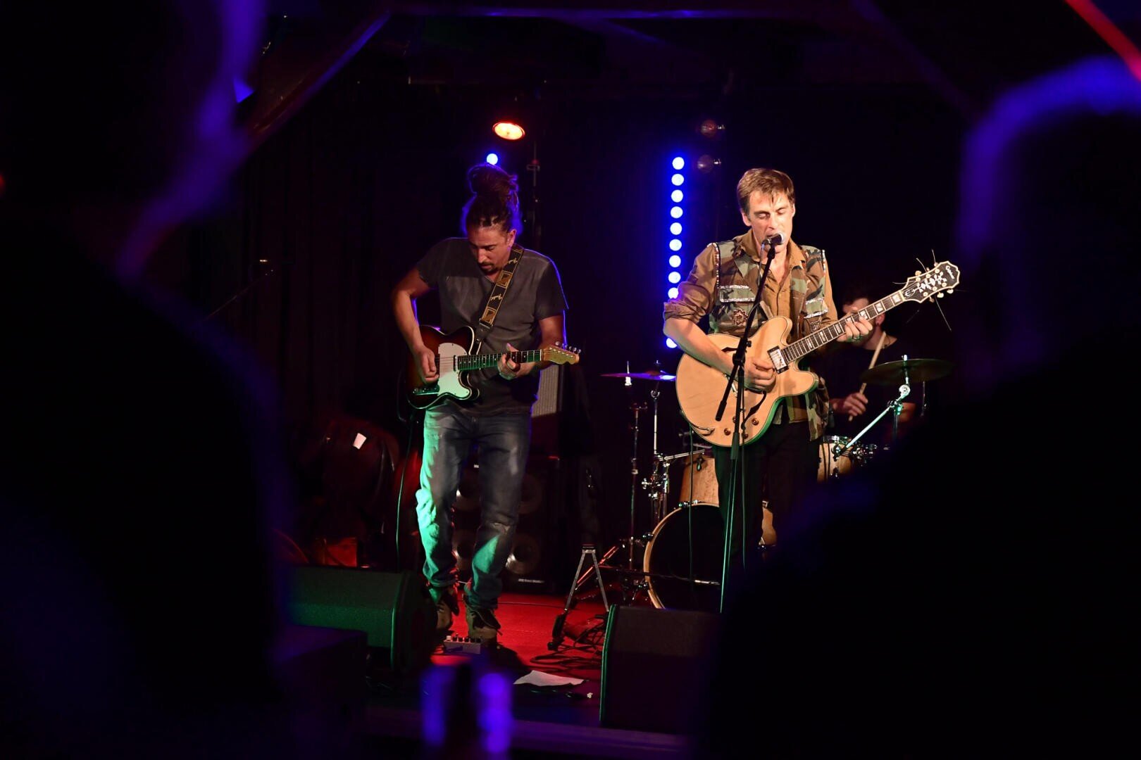 Zwei Musiker treten auf einer schwach beleuchteten Bühne auf, einer spielt elektrische Gitarre und der andere singt in ein Mikrofon, während er akustische Gitarre spielt; im Hintergrund sind Schlagzeug und Bühnenlichter zu sehen.