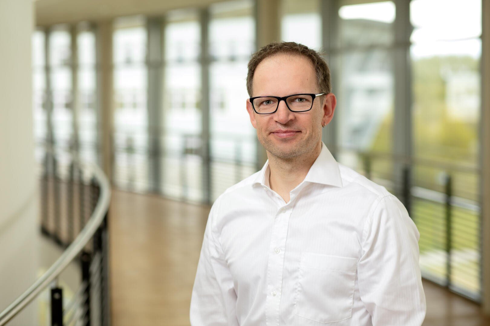 Ein Mann mit Brille und weißem Hemd steht in einem Innenraum an einem Geländer, wobei große Fenster und ein unscharfer Hintergrund natürliches Licht hereinlassen.