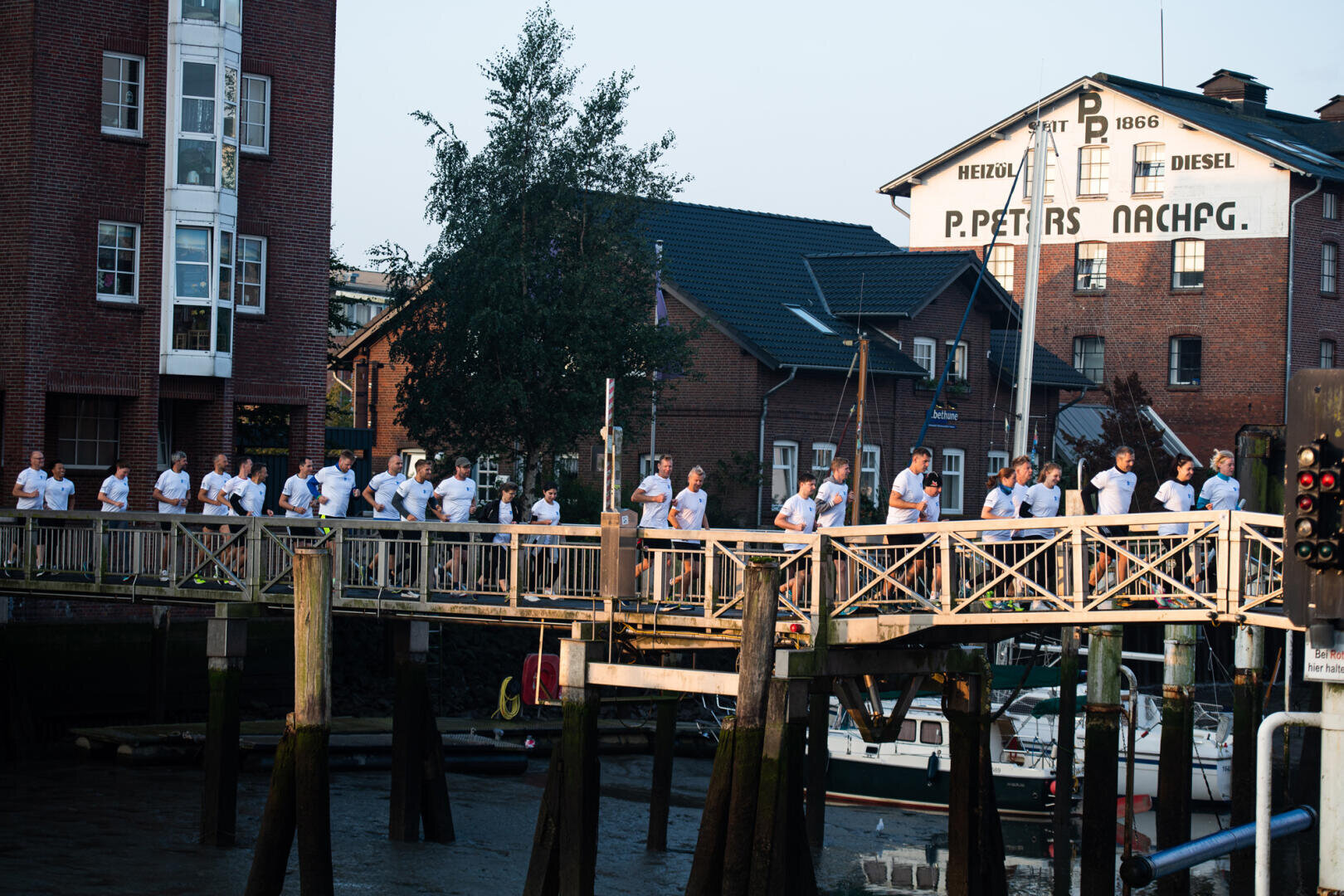A group of people in white shirts stand in a row on a wooden bridge over the water, with brick buildings and a white sign reading P. PETERS NACHFG. visible in the background.