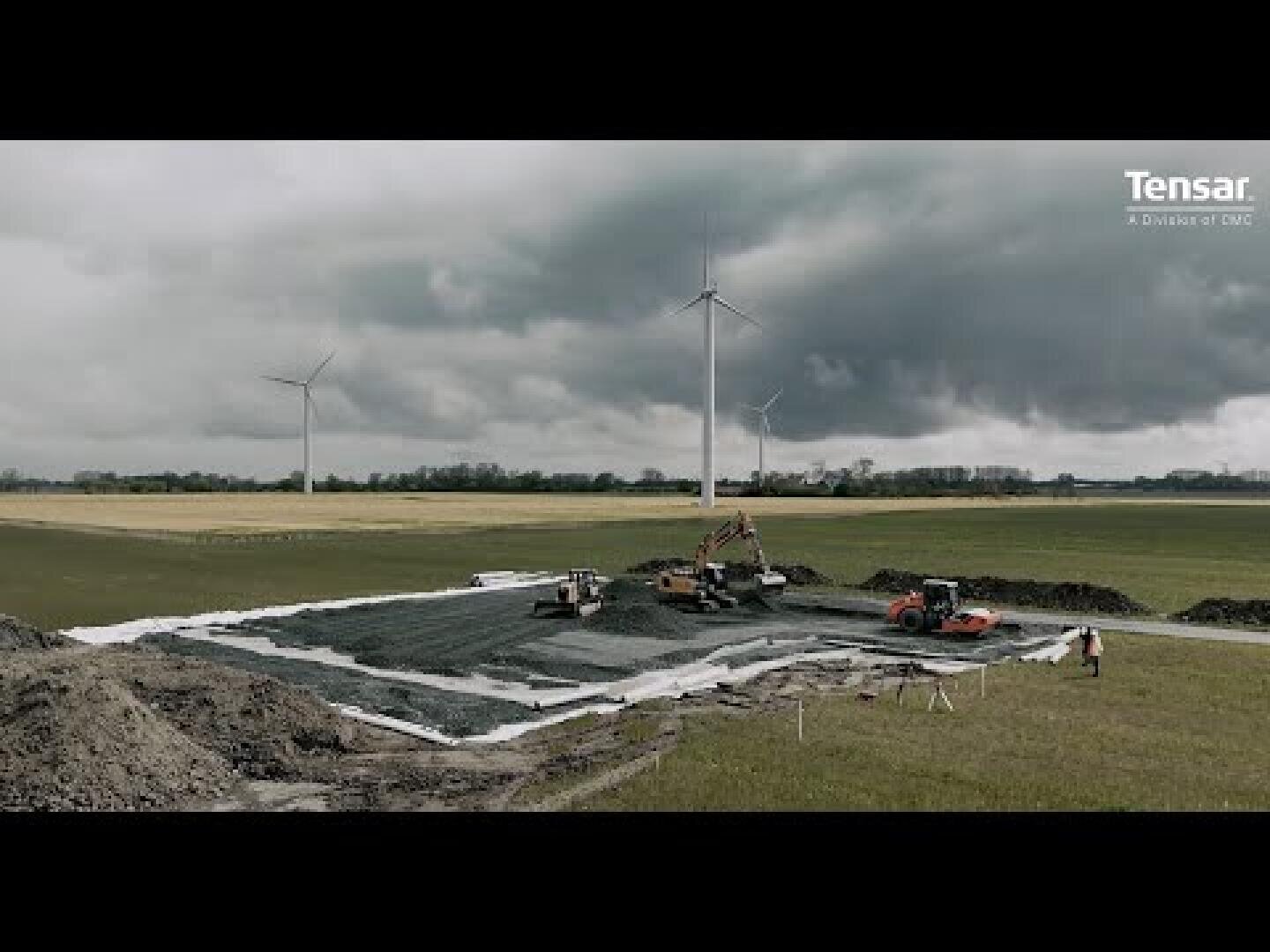 Construction vehicles work on a large rectangular area covered with geogrid material in a field, with wind turbines and cloudy skies in the background. The word “Tensar” is visible in the top-right corner.