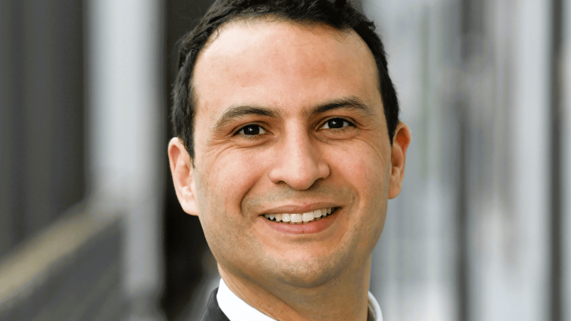 A man with short dark hair smiles at the camera. He is dressed in a suit and stands outside in front of a blurred background.