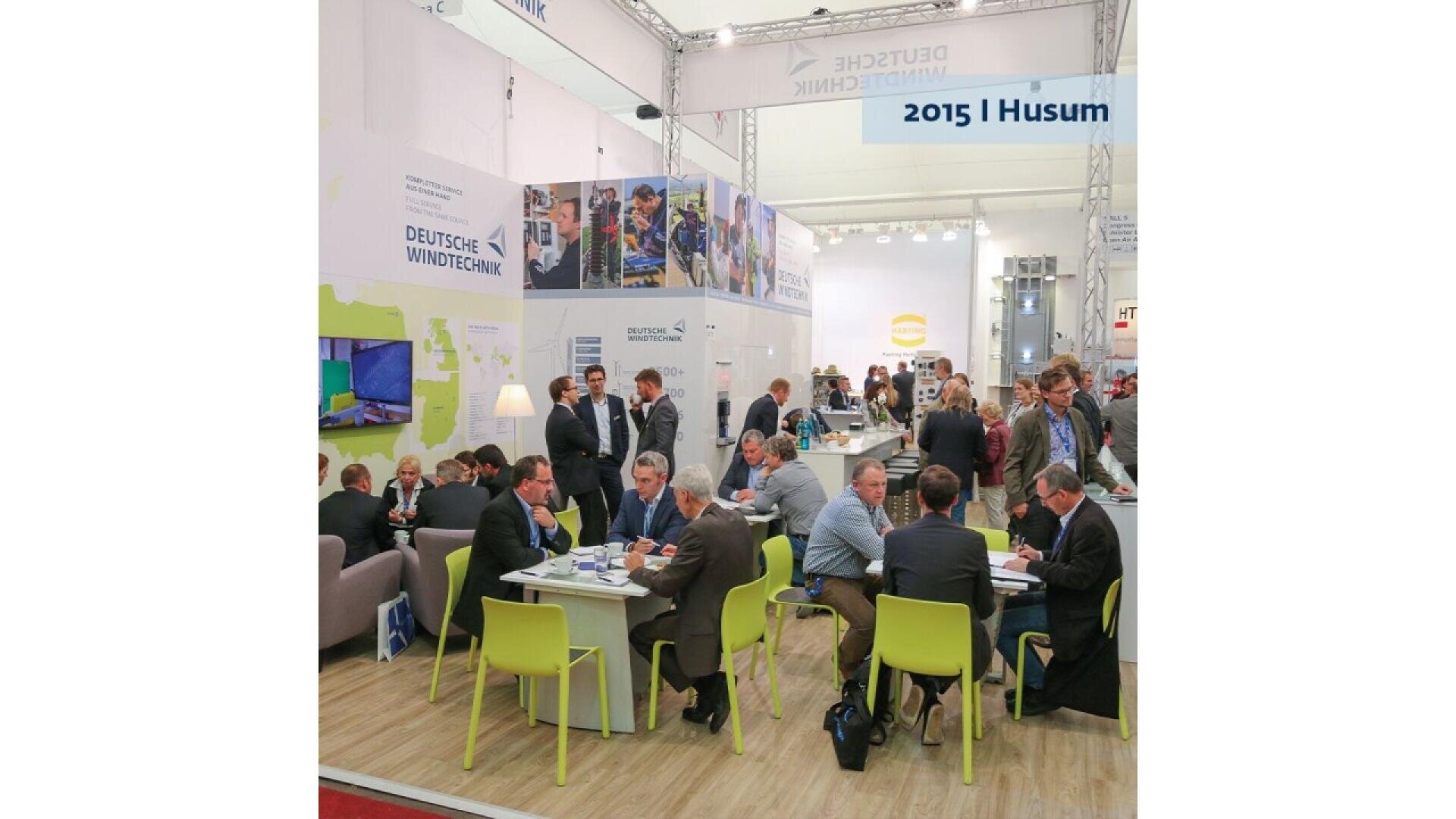People in business attire sit and talk at tables in a busy expo booth with DEUTSCHE WINDTECHNIK branding. Posters and screens are displayed on the white walls. The image is labeled 2015 | Husum.