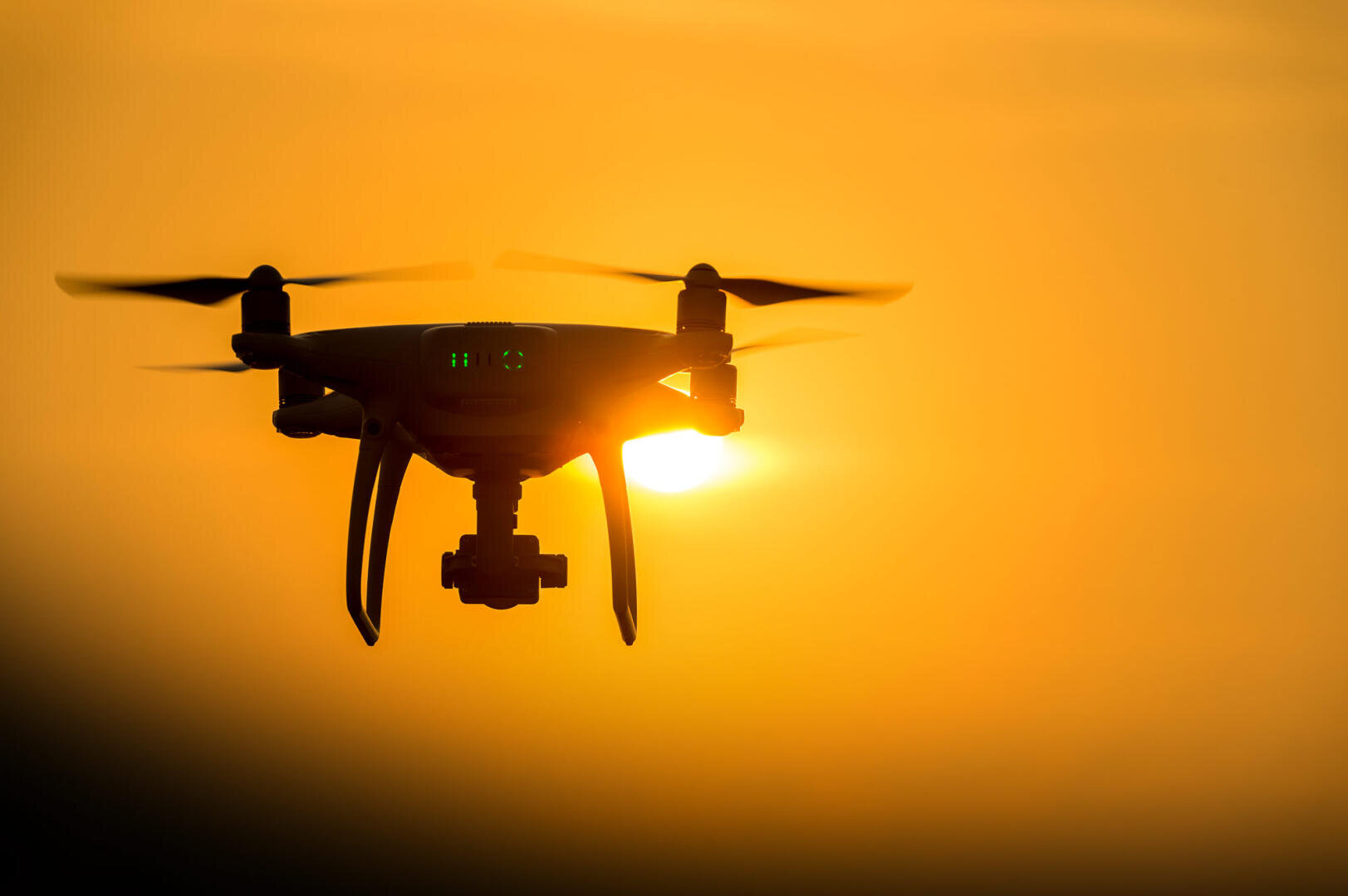 Die Silhouette einer Drohne im Flug hebt sich vor einem leuchtend orangefarbenen Abendhimmel ab, wobei die tief stehende Sonne dahinter teilweise sichtbar ist.