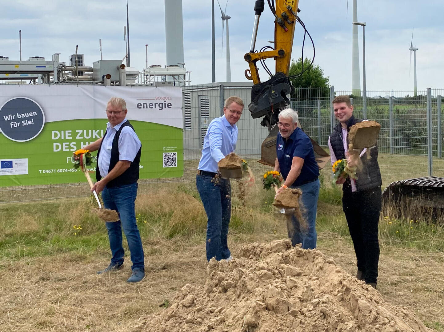 Vier Männer mit Schaufeln brechen den Boden auf einer Baustelle in der Nähe von Windkraftanlagen auf und werfen Sand in die Luft. Im Hintergrund sind eine große Baumaschine und ein Banner in deutscher Sprache zu sehen.