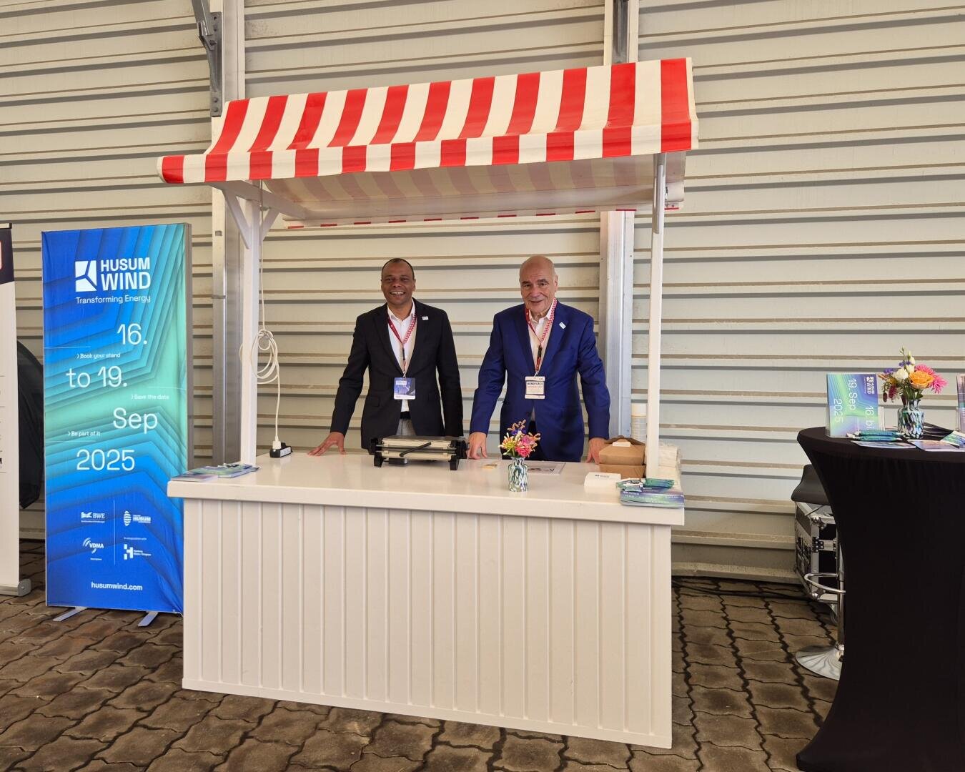 Zwei Männer in Anzügen stehen lächelnd hinter einem weißen Stand mit einem rot-weiß gestreiften Vordach. Ein blaues Husum Wind Veranstaltungsbanner und ein schwarzer Tisch mit Broschüren stehen neben ihnen in einem Ausstellungsraum.