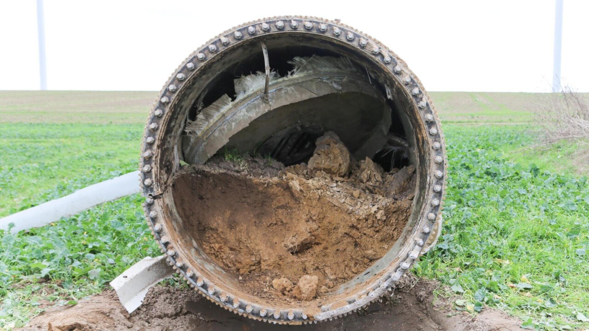 A large, broken wind turbine blade lies horizontally on green grass, with the interior filled with dirt and debris, exposing its damaged, fibrous structure.