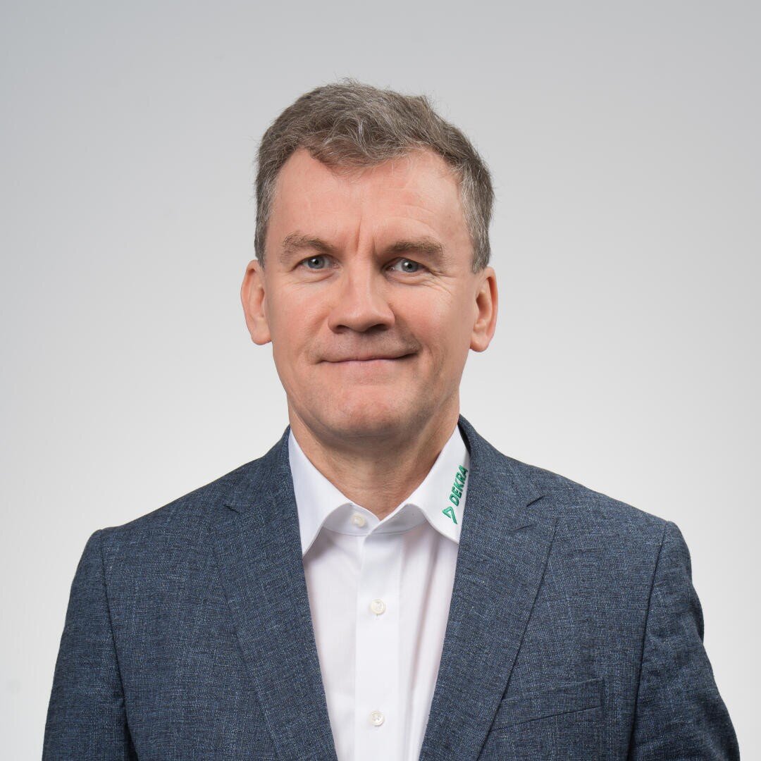 A man with short brown hair, wearing a blue blazer and a white collared shirt, stands in front of a plain light gray background and smiles slightly.