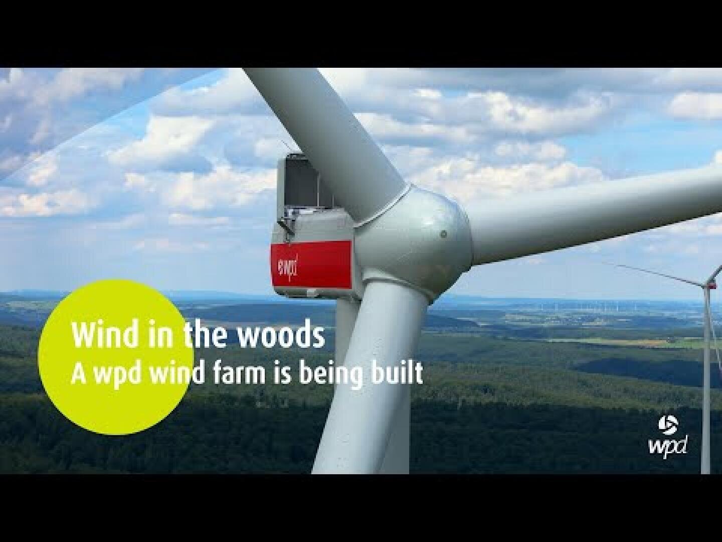 Close-up of a wind turbine with a forest and hills in the background. Text reads, Wind in the woods. A wpd wind farm is being built. The wpd logo appears in the bottom right corner.