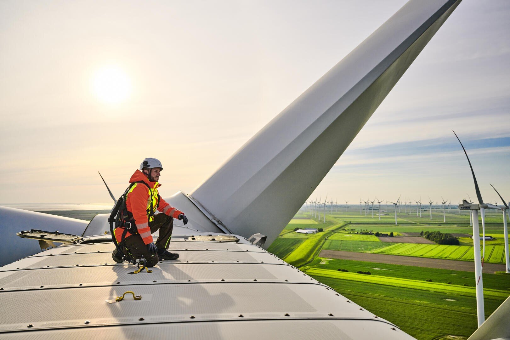 Ein Arbeiter in Schutzkleidung sitzt auf einer Windturbine und überblickt grüne Felder und andere Turbinen unter einem hellen Himmel, während die Sonne tief am Horizont steht.
