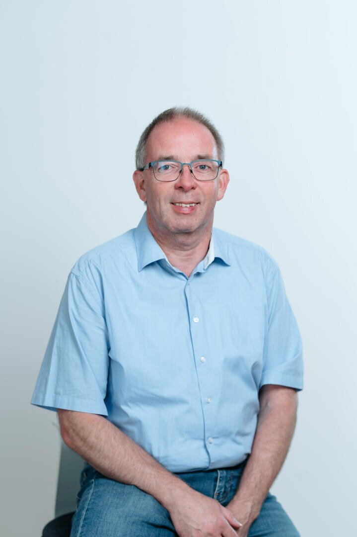 A middle-aged man with short gray hair and glasses is sitting on a chair, wearing a light blue short-sleeved shirt and blue jeans, against a plain light background. He is smiling slightly and looking at the camera.