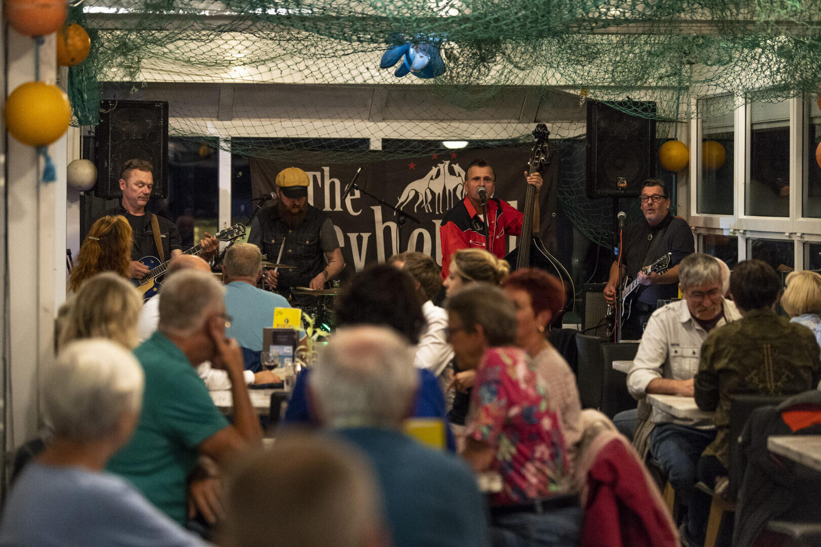 Eine Band spielt Livemusik auf einer kleinen, mit Fischernetzen dekorierten Bühne im Innenbereich, während eine Menge sitzender Menschen zuschaut und an Tischen in einem zwanglosen, geschäftigen Restaurantambiente speist.