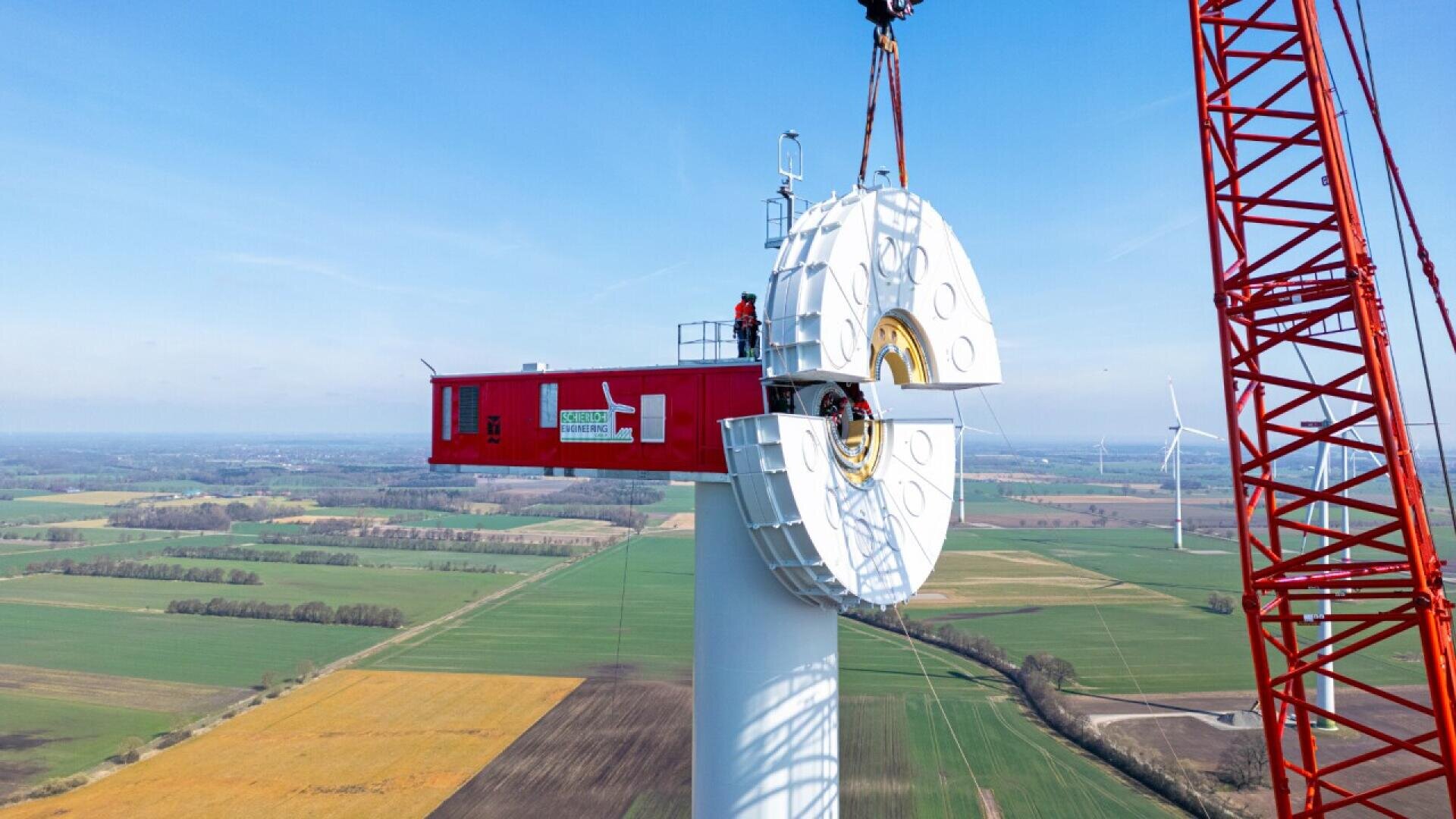 A large red crane lifts a wind turbine nacelle onto the top of a tall wind turbine tower in a rural landscape, with workers visible on the structure and other wind turbines in the background.