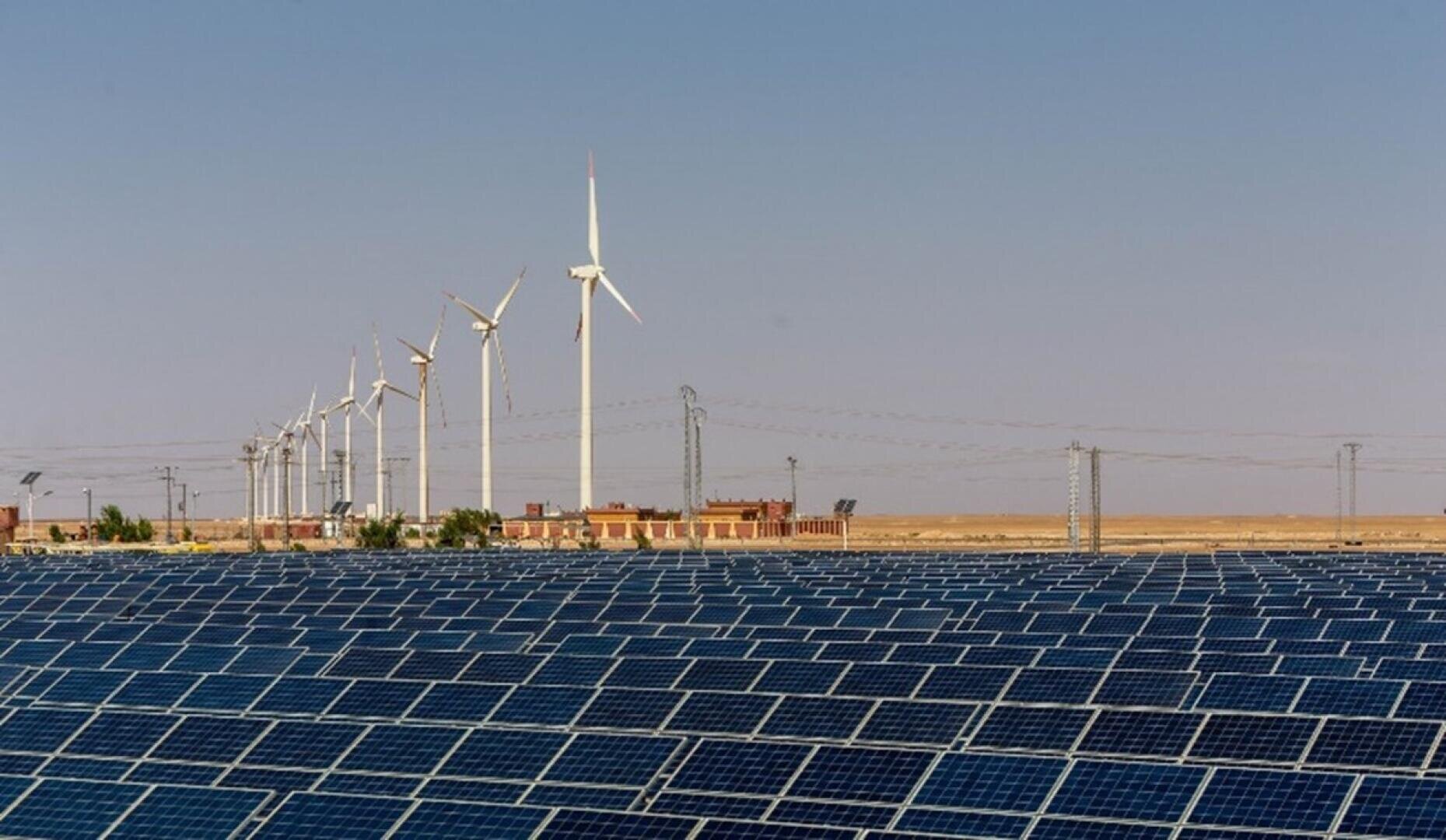 Reihen von Solarmodulen bedecken den Vordergrund, während im Hintergrund mehrere Windturbinen vor einem klaren blauen Himmel und einer flachen, trockenen Landschaft stehen. Auch kleine Gebäude und Stromleitungen sind zu sehen.